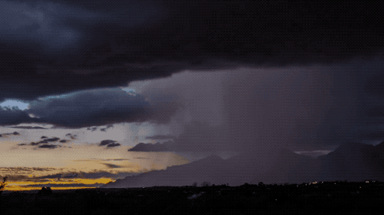 This animated GIF shows clouds moving across a dusky sky. The clouds on the right side have a gray haze extending down to the bottom of the image, where there is rain. Flashes of lightning, stretching from the cloud to the ground, light up the screen periodically.
