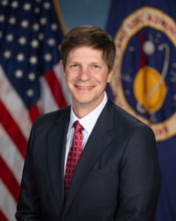 Portrait of Kevin Murphy, male, in front of a United States flag and a NASA seal flag wearing a black suit jacket and red tie.