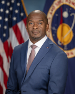 Portrait of Andrew Mitchell, male, in front of a United States flag and a NASA seal flag wearing a blue suit jacket and red tie.