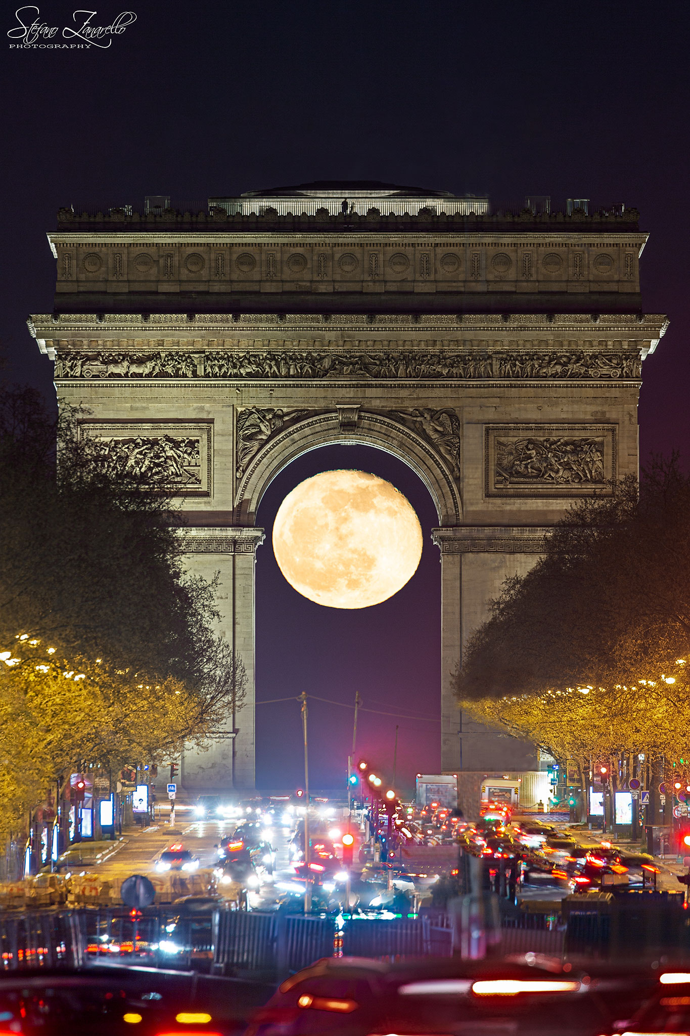 A nearly full Moon is seen through the famous Arc de Triomphi with trees and cars lining the foreground. Please see the explanation for more detailed information.