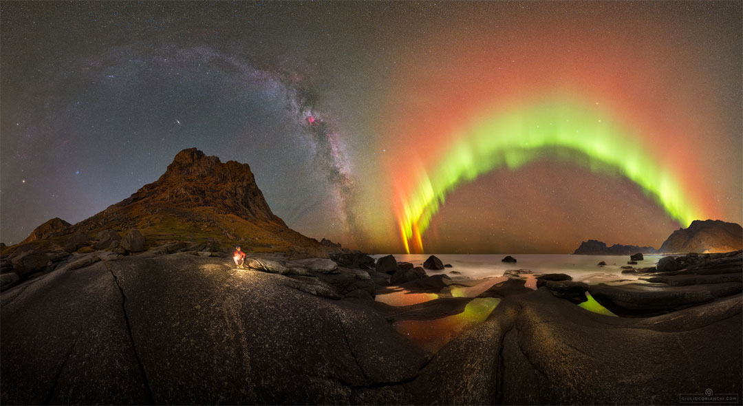 A night sky filled with stars is shown behind a picturesque foreground. The foreground contains rounded rocks and a person before a distant sea. The background contains bands of the Milky Way and bright aurora. Please see the explanation for more detailed information.