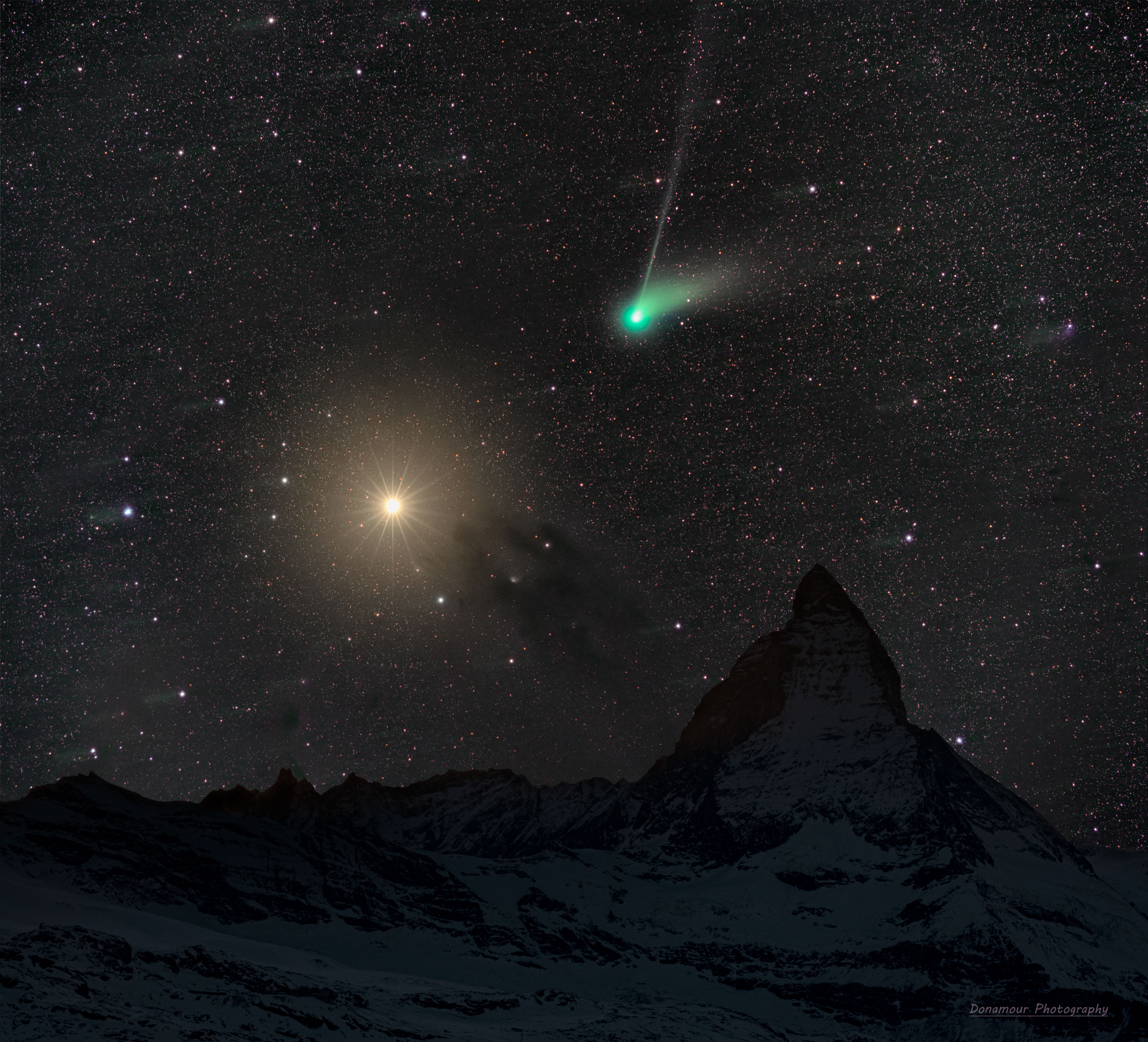 A deep starfield features an orange planet Mars on the left and a green-headed Comet ZTF on the right. In the foreground is a landscape that includes the top of a famous mountain known as the Matterhorn. Please see the explanation for more detailed information.