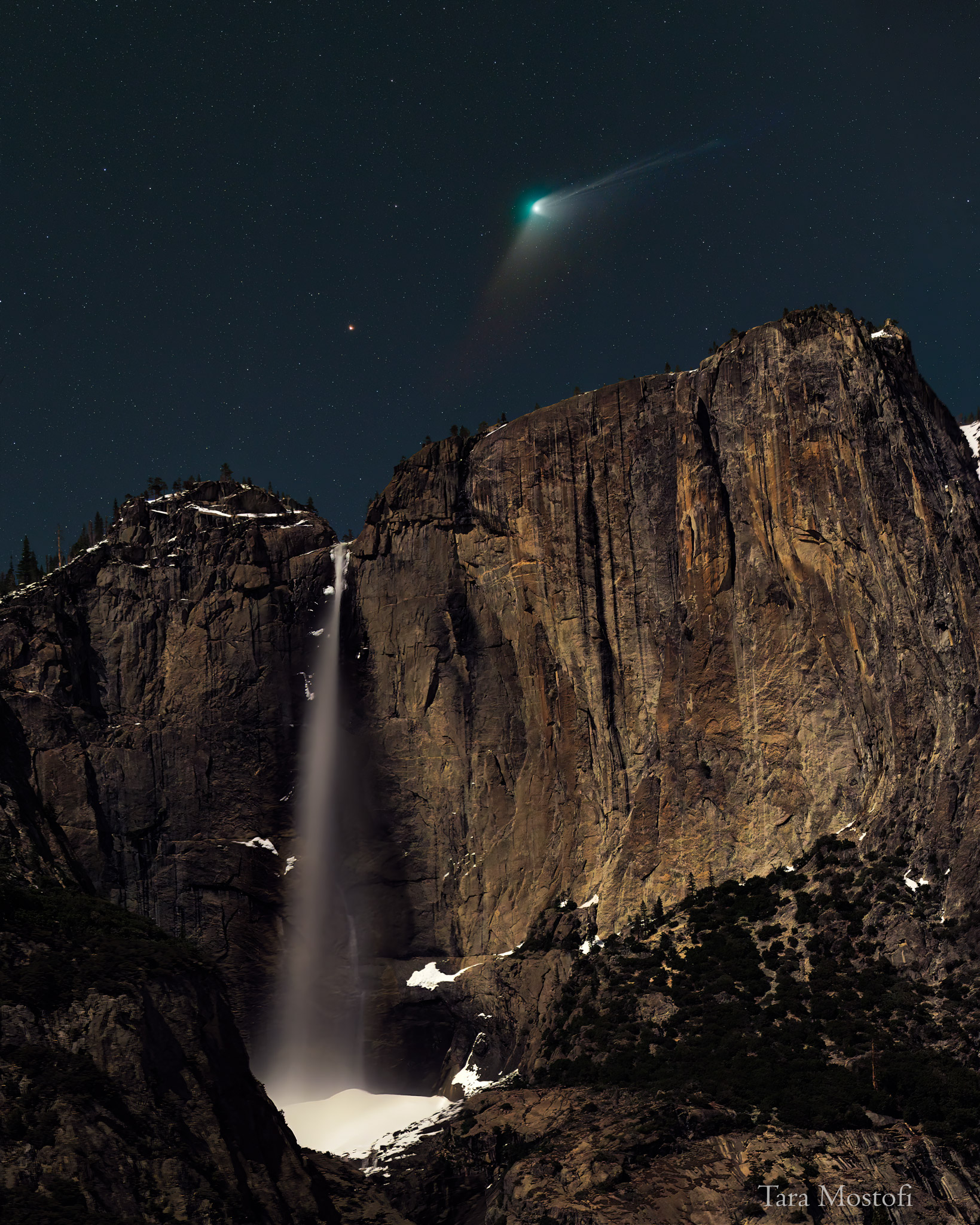 A comet with a green head and extended tails is seen above a high water fall. In the night sky field just above the falls, an orange dot -- the star Kochab -- is visible. Please see the explanation for more detailed information.