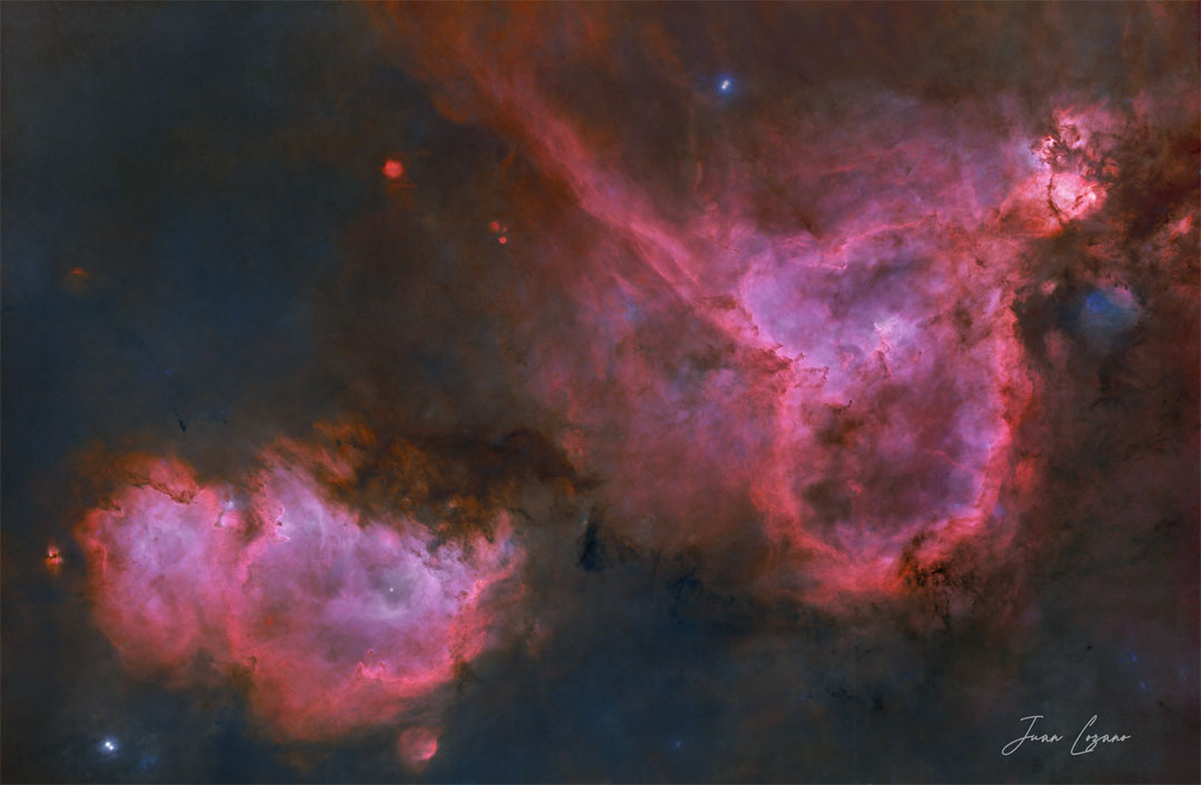 Two red emission nebulas are shown in front of a dark but colorful starfield. The Soul Nebula is on the lower left, while the Heart Nebula is on the upper right. Please see the explanation for more detailed information.