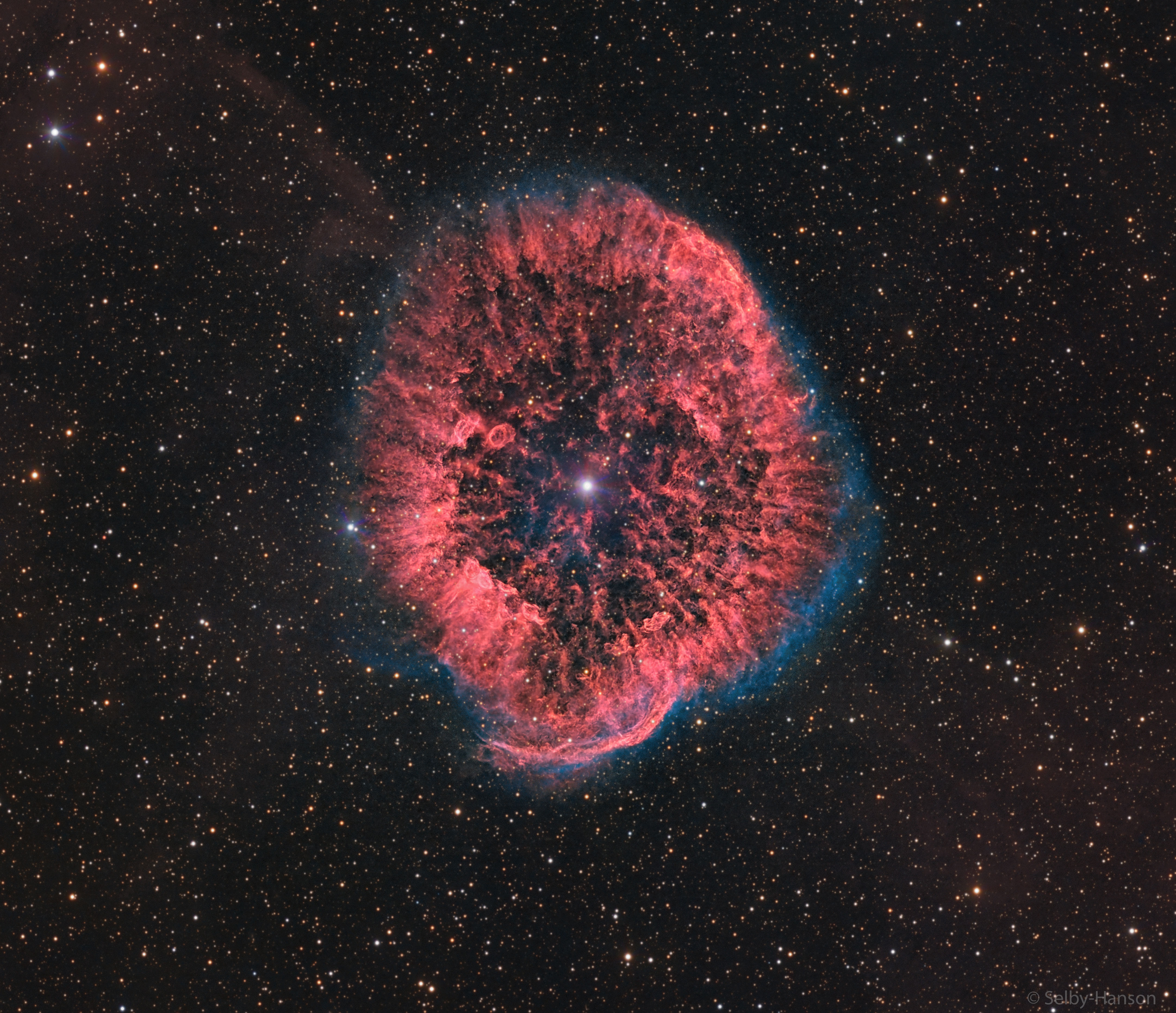 A red oval and textured nebula is seen surrounded by a faint blue glow. A bright star is visible in the center, and many faint stars are visible in the background. Please see the explanation for more detailed information.