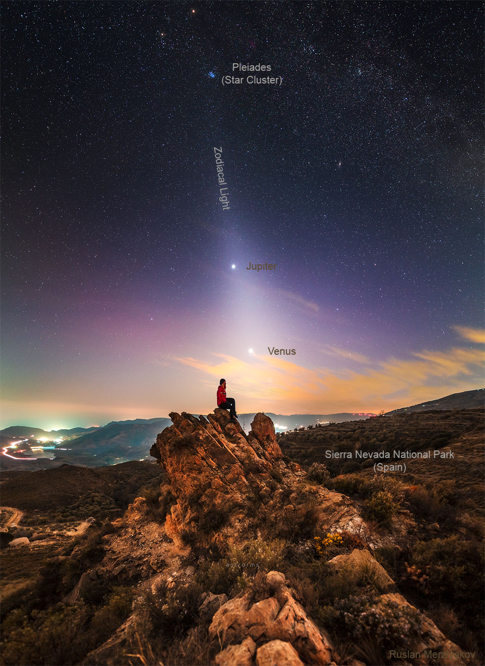 A person is seen sitting on a rock under an unusual sky. In the sky above is light diffuse band extending down to the horizon that goes through two bright dots, Jupiter and Venus. The Pleiades star cluster is visible above them. Please see the explanation for more detailed information.
