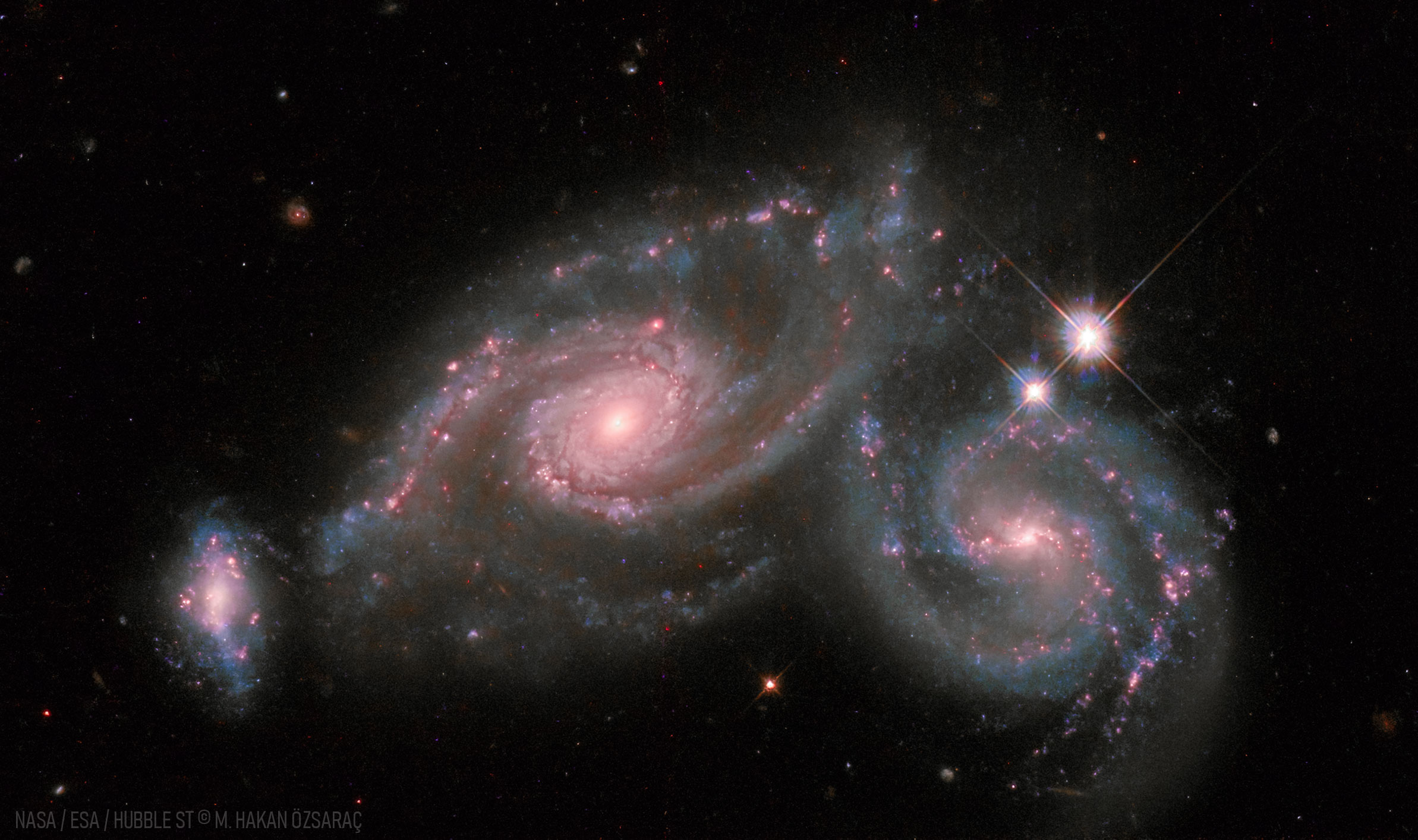 Two spiral galaxies are shown right next to each other, with a smaller distorted galaxy on the far left. Please see the explanation for more detailed information.