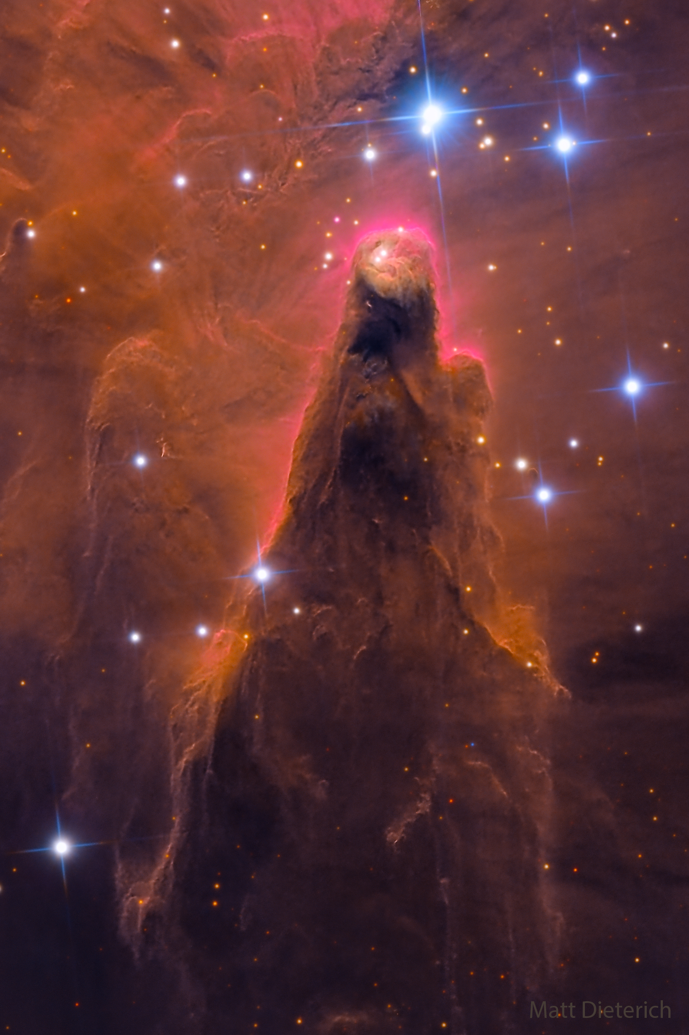 A conical interstellar dust pillar is pictured. The pillar is mostly brownish-red but surrounded by stars. Please see the explanation for more detailed information.