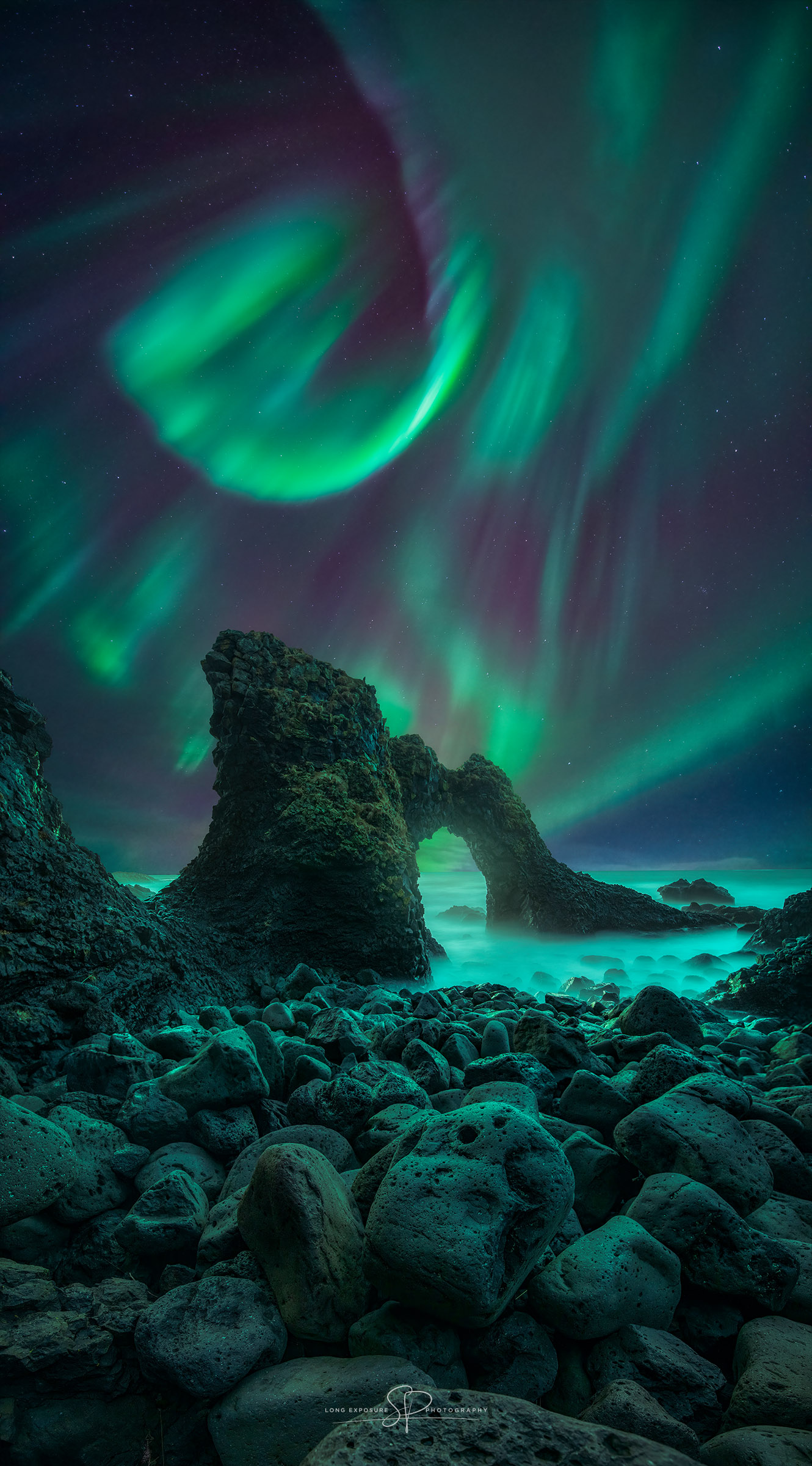 A green aurora is pictured above and beyond a dark rocky arch. Faint stars dot the background. Please see the explanation for more detailed information.