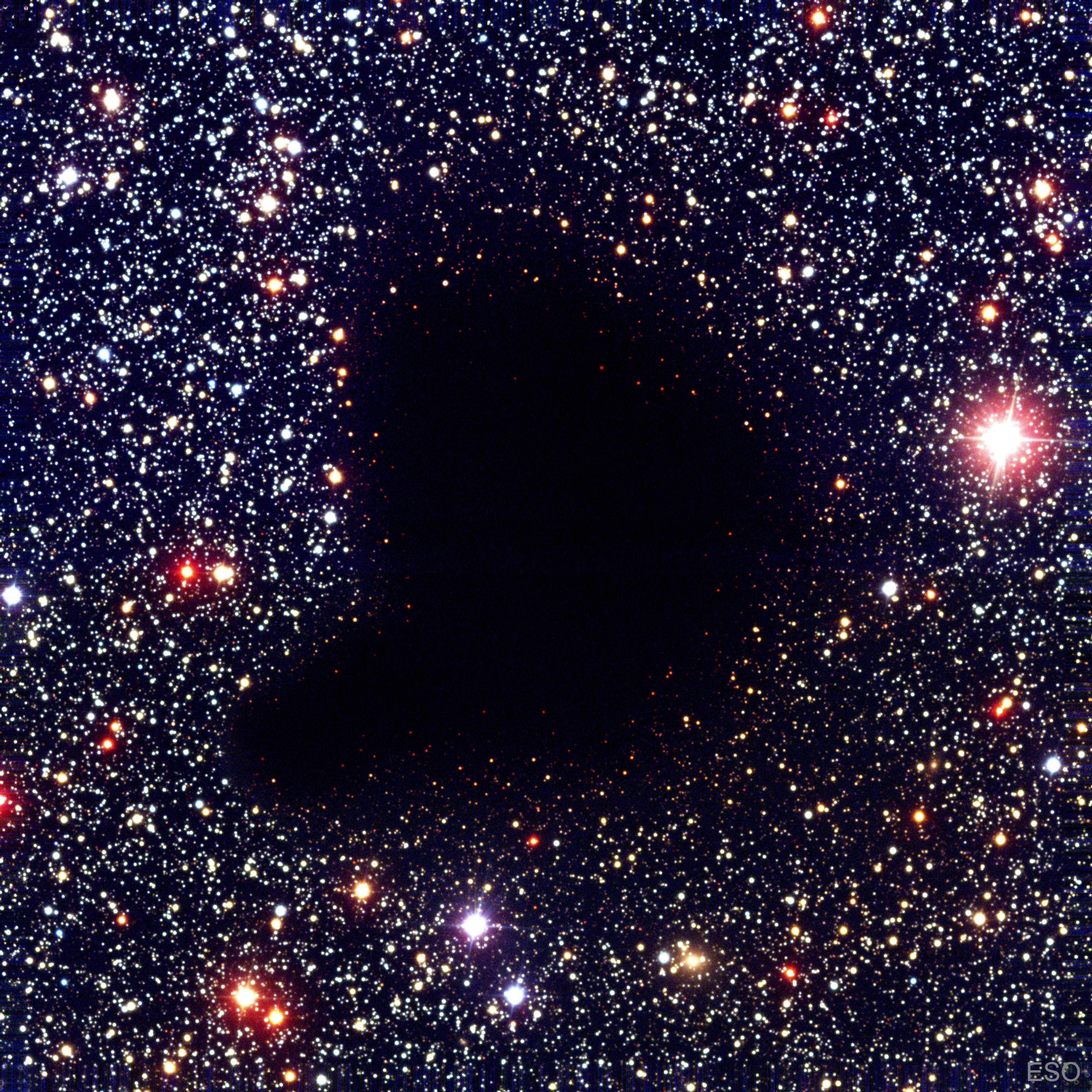 A dark comma-shaped cloud appears in the middle of a dense field of stars. No stars are visible through the center of the cloud. Please see the explanation for more detailed information.