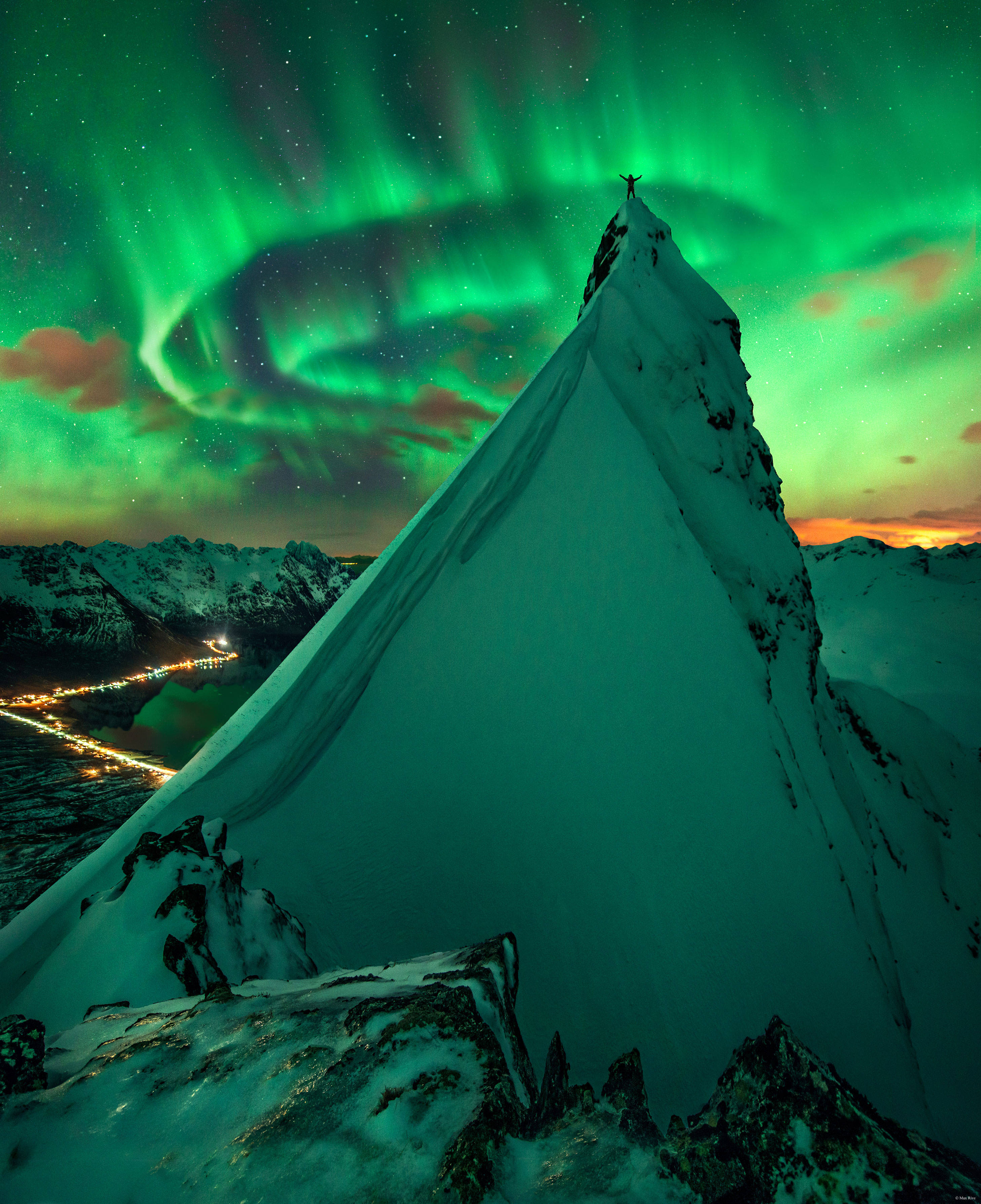 A person stands on a steep snow-covered hill with their arms raised. In the distance green aurora are visible. Past that stars are visible. Please see the explanation for more detailed information.