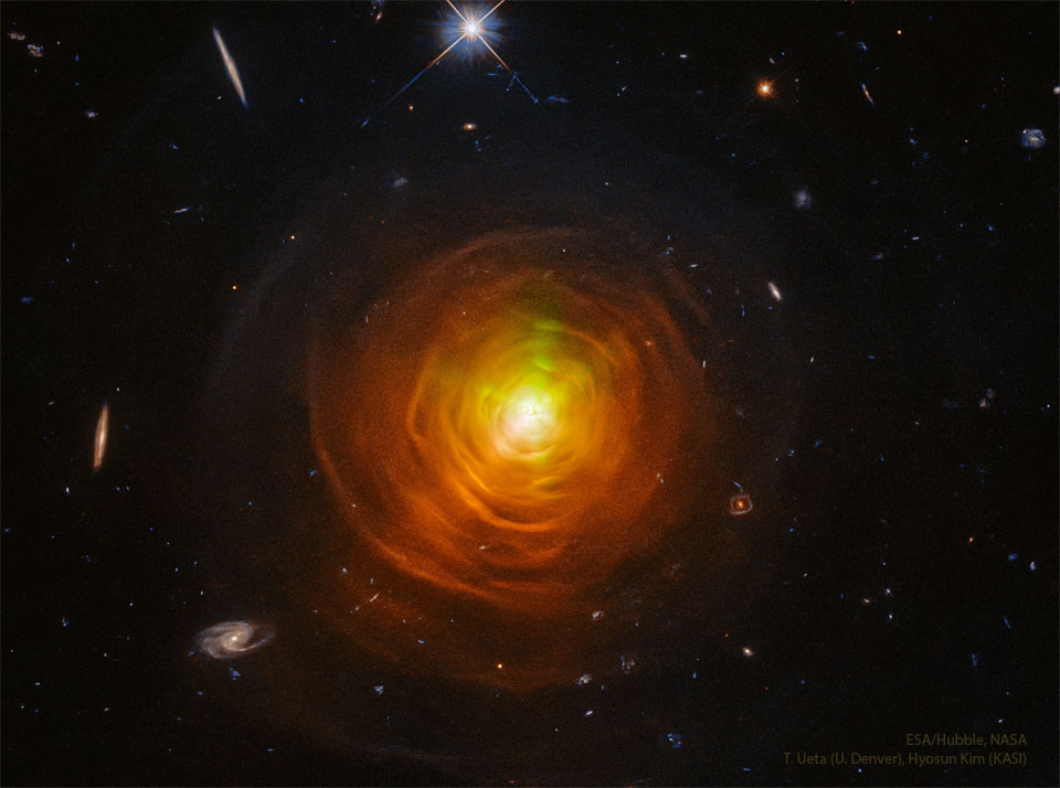 A star surrounded by orange shells and arcs sit in the center of a dark starfield. Galaxies from the distant universe can be seen around the edges. Please see the explanation for more detailed information.