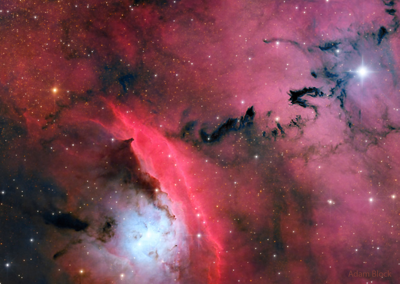 A busy star formation region is shown highlighted by red glowing clouds and dark ominously-shaped dust. Please see the explanation for more detailed information.