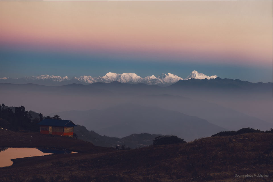An orange sky hovers above snow-covered mountains. A blurry line divides the orange sky from a darker sky. In the foreground are hills and a house. Please see the explanation for more detailed information.