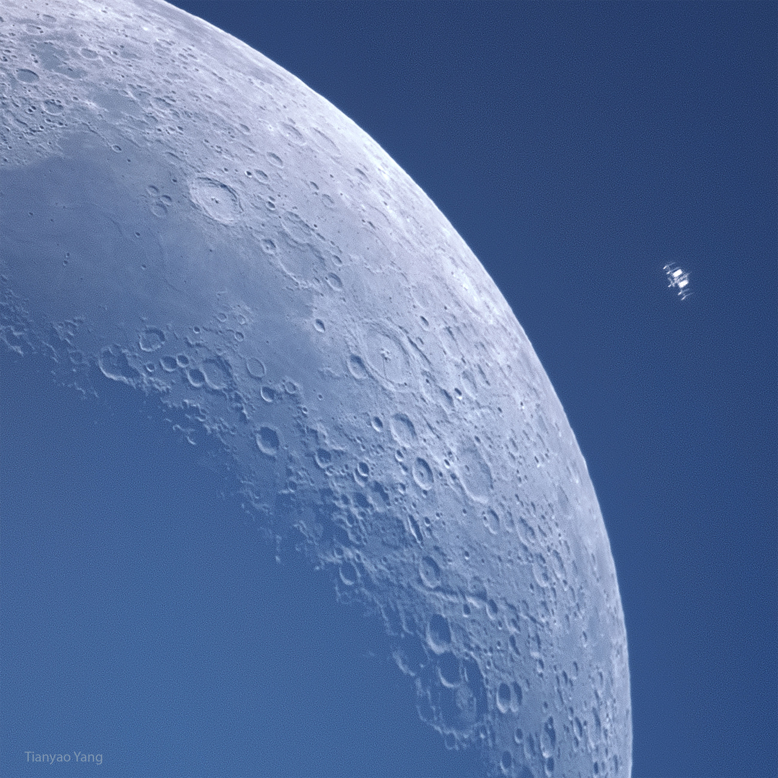 A crescent moon is shown against blue background. Many craters are visible in great detail. To the upper left appears some kind of small machine which is actually the International Space Station also in orbit around the Earth. Please see the explanation for more detailed information.