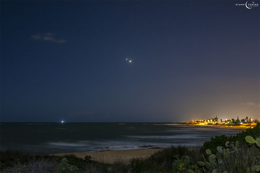 Two bright objects are pictured very near each other in night sky filled with stars. A beach is in the foreground, with some lit structures visible across the water. Please see the explanation for more detailed information.