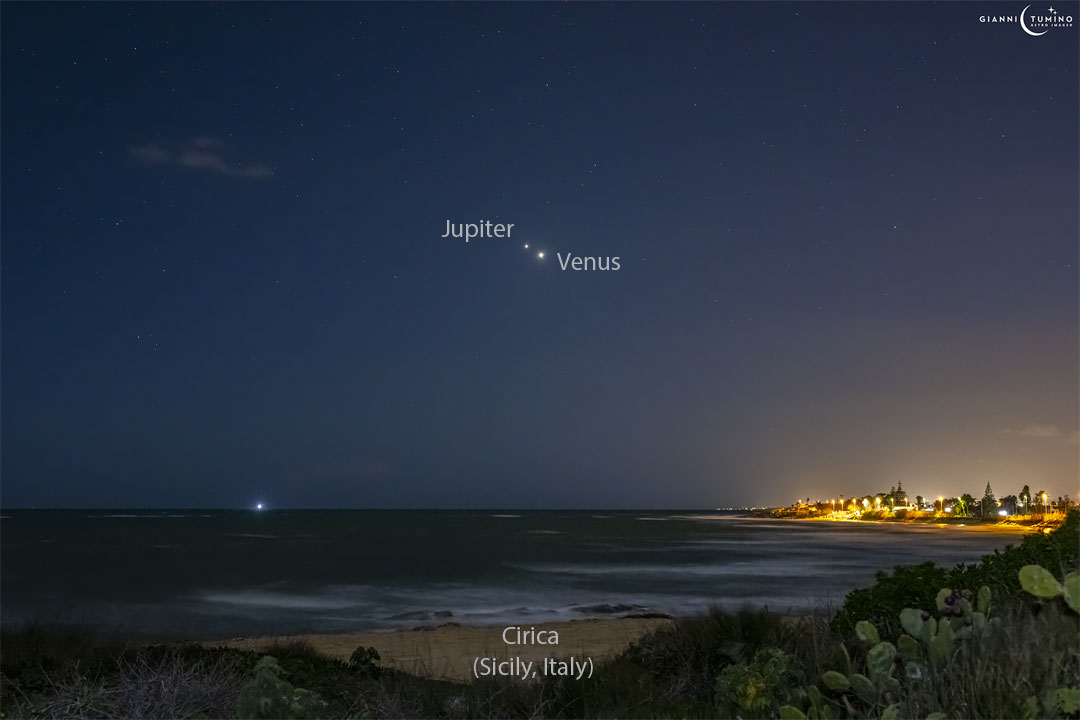 Two bright objects are pictured very near each other in night sky filled with stars. A beach is in the foreground, with some lit structures visible across the water. Please see the explanation for more detailed information.