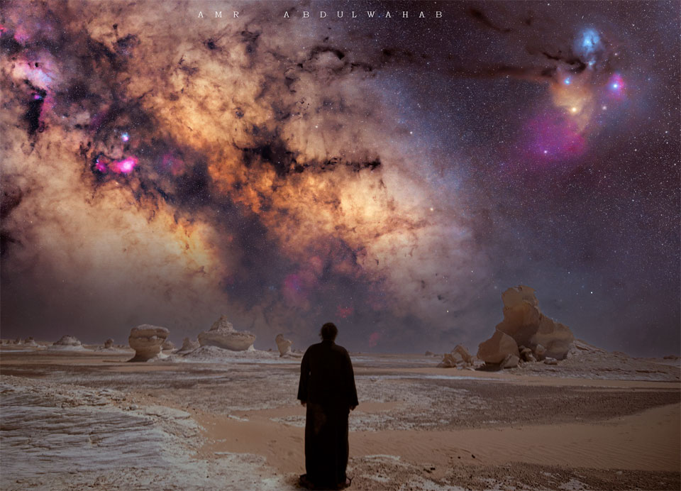 A person in silhouette looks out over a desert punctuated by unusual rock formations. High above is a colorful sky including the band of our Milky Way Galaxy and the Rho Ophiuchi star clouds. Please see the explanation for more detailed information.
