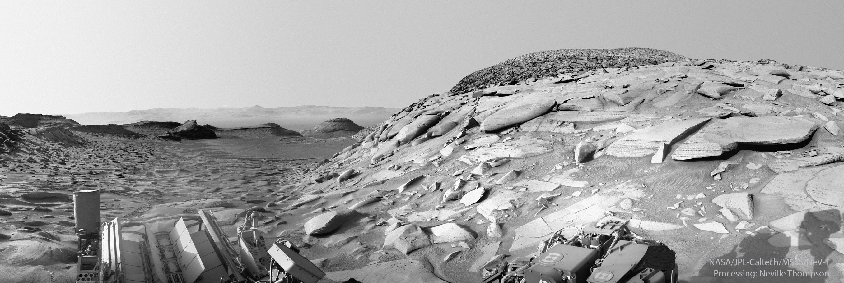 A view of Mars from the Curiosity rover on Mars is pictured in black and white. Many rocks and hills are visible, with a hill containing many unusually flat rocks visible on the right. Please see the explanation for more detailed information.