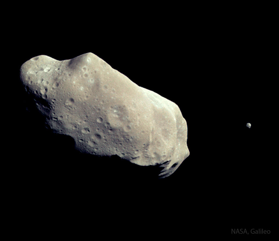 A pair of asteroids are shown with a large, elongated and cratered one on the left and a much smaller one on the far right. Please see the explanation for more detailed information.