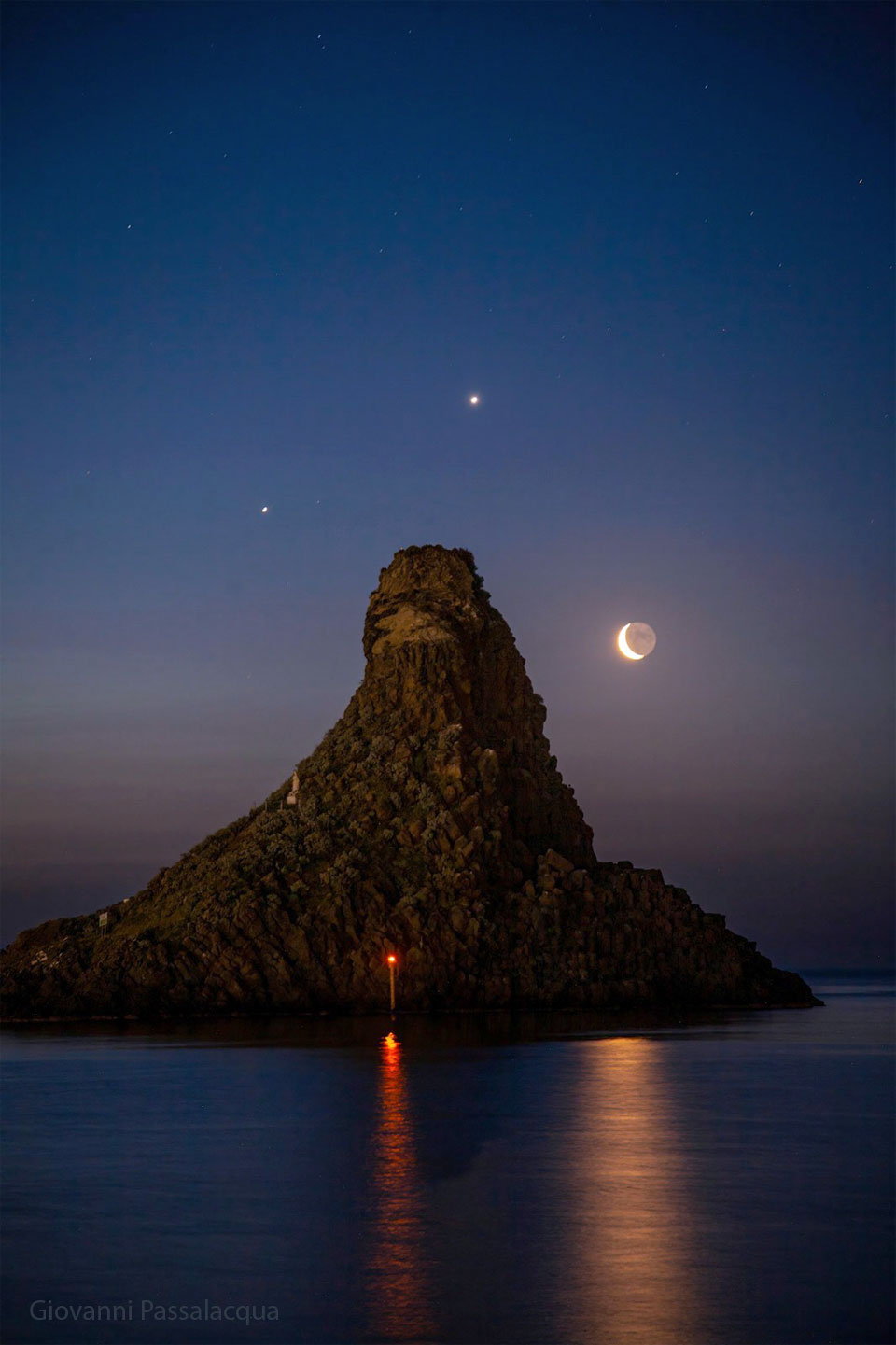 A seascape surrounds a large tree-covered hill. Surrounding the hill in the night sky are three bright dots: the planets Jupiter, Venus, and a crescent Moon. Please see the explanation for more detailed information.