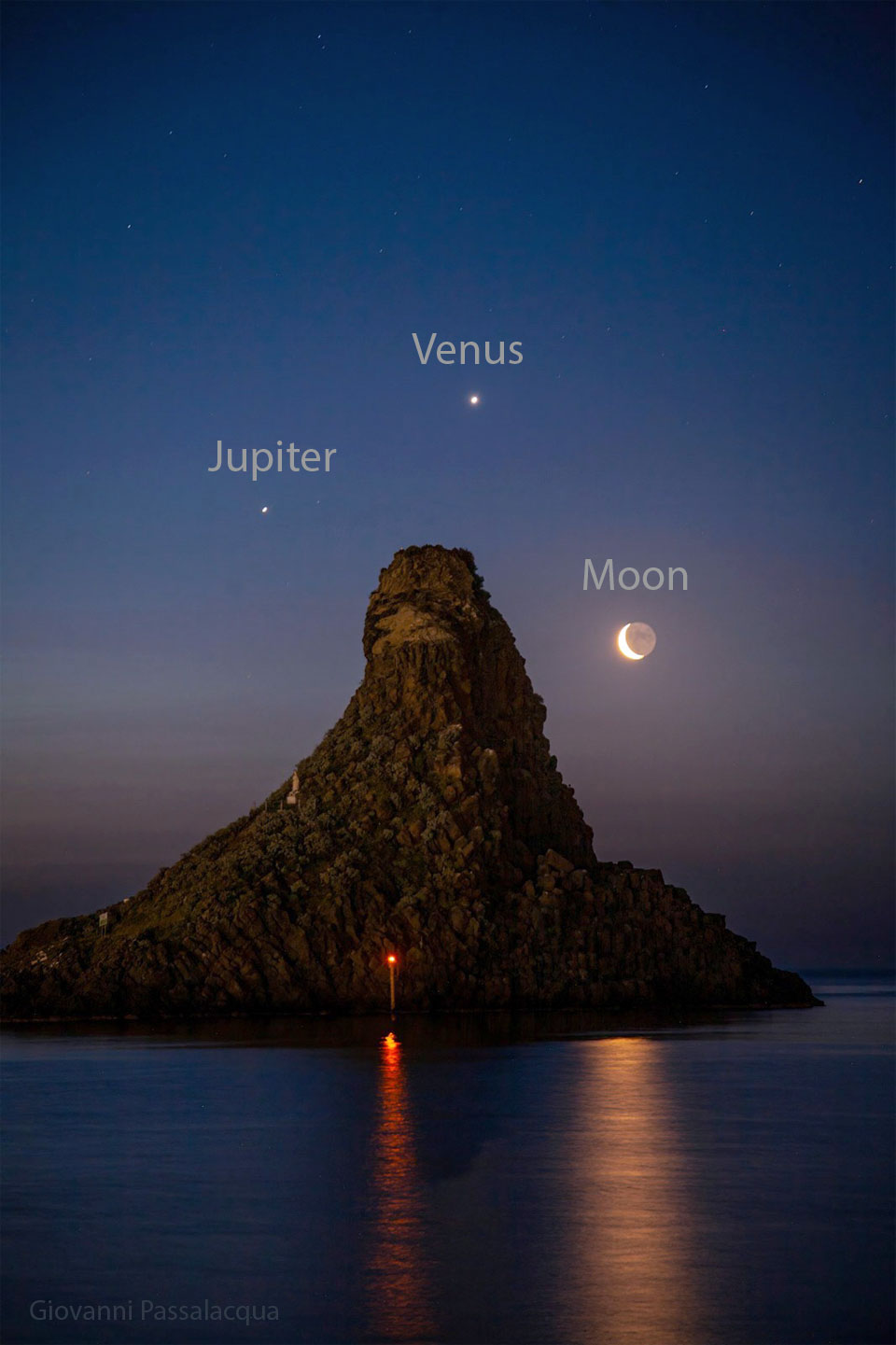 A seascape surrounds a large tree-covered hill. Surrounding the hill in the night sky are three bright dots: the planets Jupiter, Venus, and a crescent Moon. Please see the explanation for more detailed information.