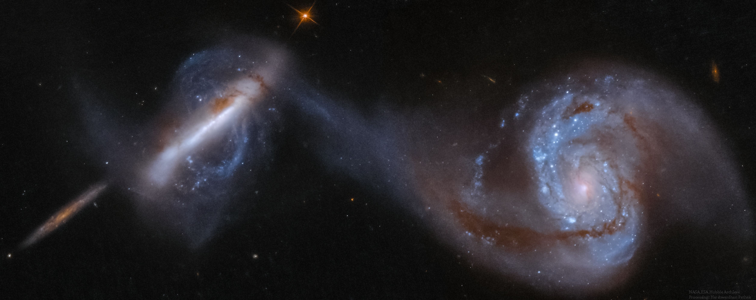 Three large galaxies are shown, the rightmost two in collision. The galaxy on the far right is a large spiral galaxy with one arm connected to an unusual polar galaxy on the left. The smaller galaxy on the far left is thought to be far in the background. Please see the explanation for more detailed information.
