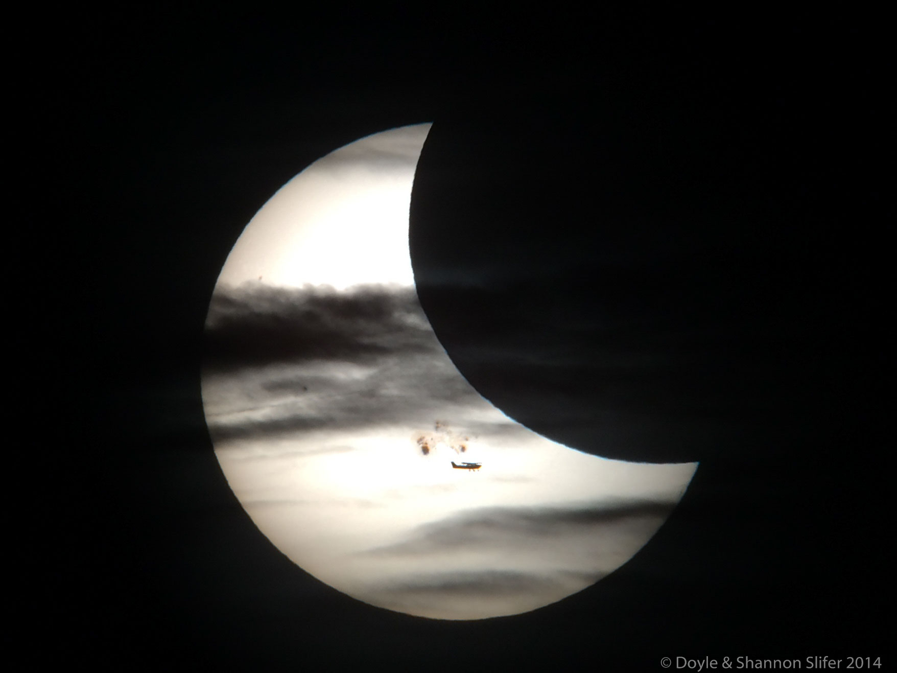 A partially eclipse Sun is shown. In front of the Sun are sunspots, the Moon, clouds, and an airplane. Please see the explanation for more detailed information.