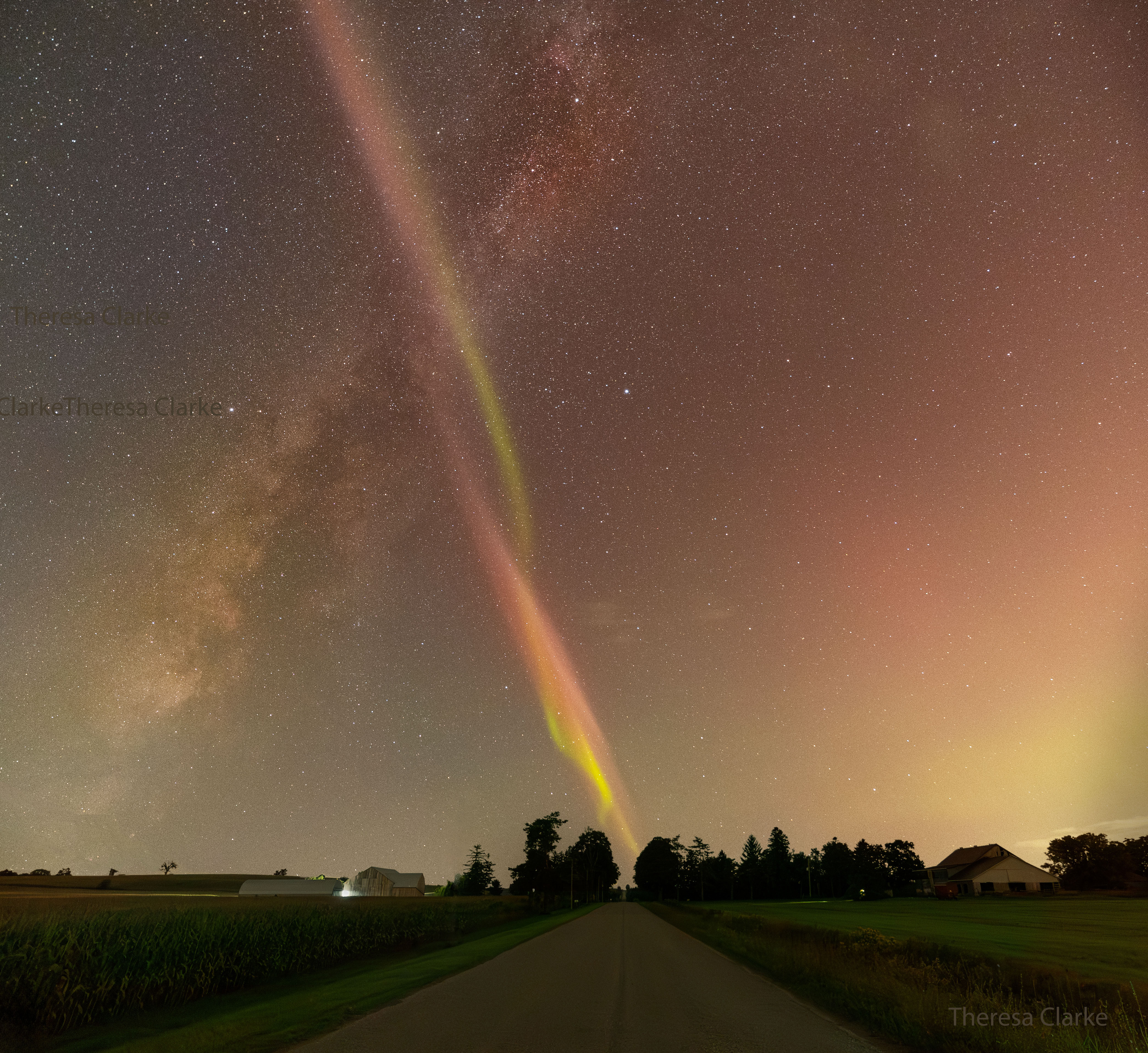 A rural road is pictured running to the horizon with rural grassy fields on both sides. Rising from the lower left is the central band of our Milky Way Galaxy. Rising from the horizon -- just at the visible end of the road, is a thin twisting band of light twisting green and red bands -- a STEVE. The STEVE crosses in front of the Milky Way band making a big