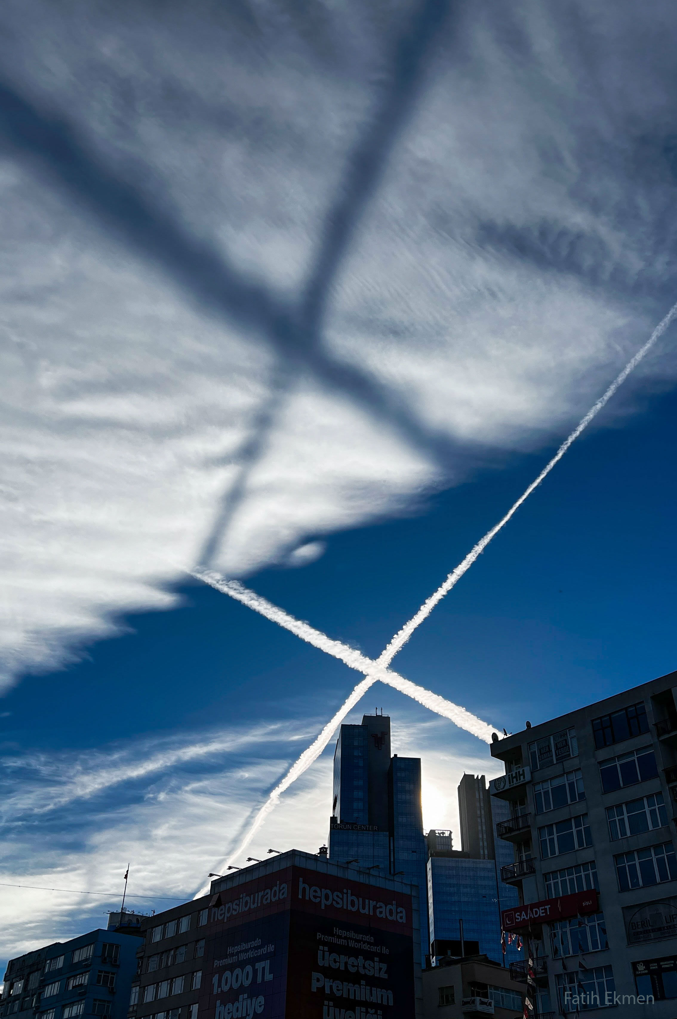 Two airplane contrails, crossing in an X, are shown across the middle of the image. They are bright white against a dark blue background. A high cloud deck is seen above the crossing, sunlit, contrails. A low Sun creates a dark shadow X on the high while clouds. A row of buildings runs across the lower part of the image. Please see the explanation for more detailed information.