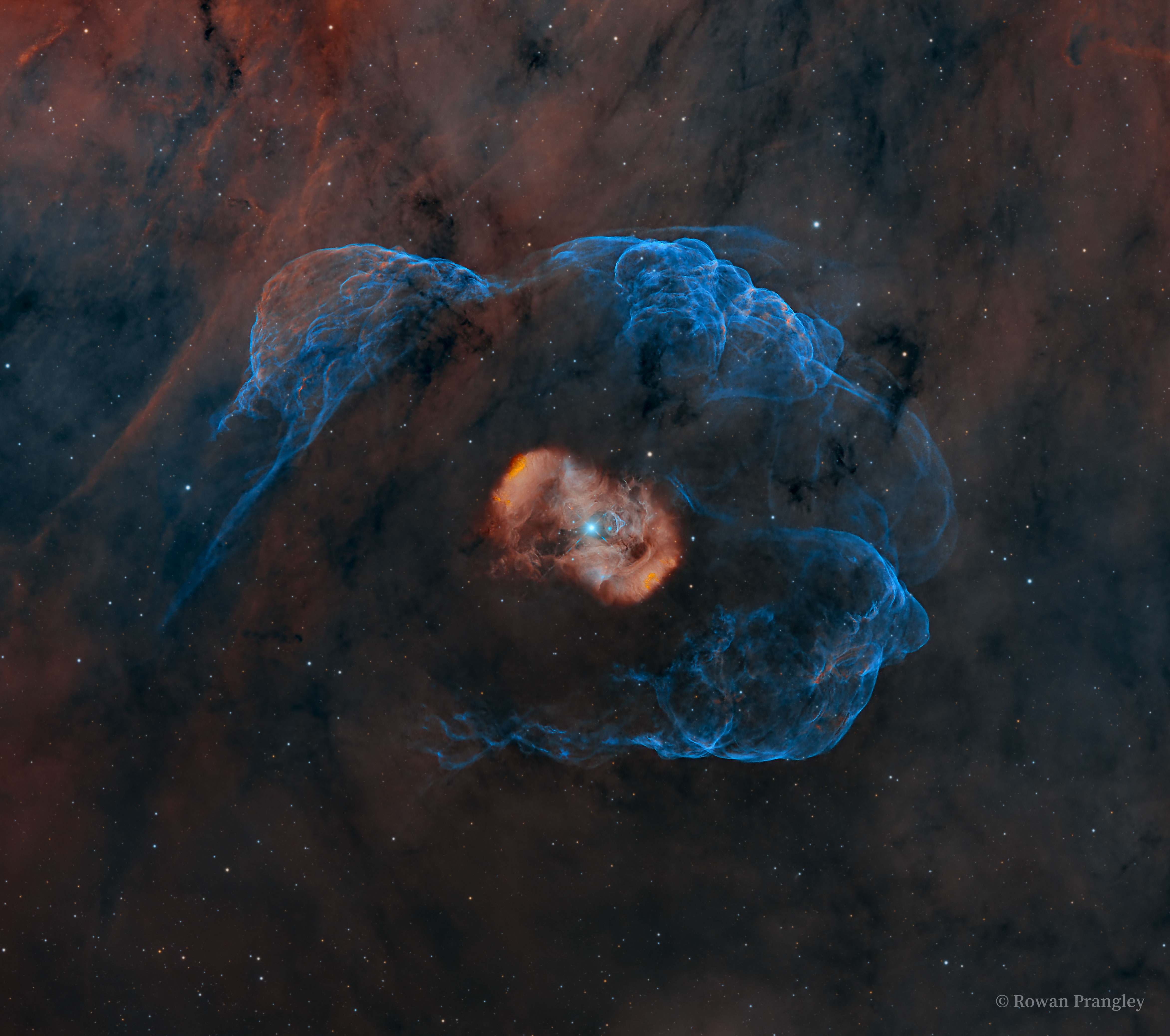 A blue star is seen in the center of a red nebula itself surrounded by a faint blue nebula. The surrounding starfield itself has a faint red-brown emission clouds. Please see the explanation for more detailed information.