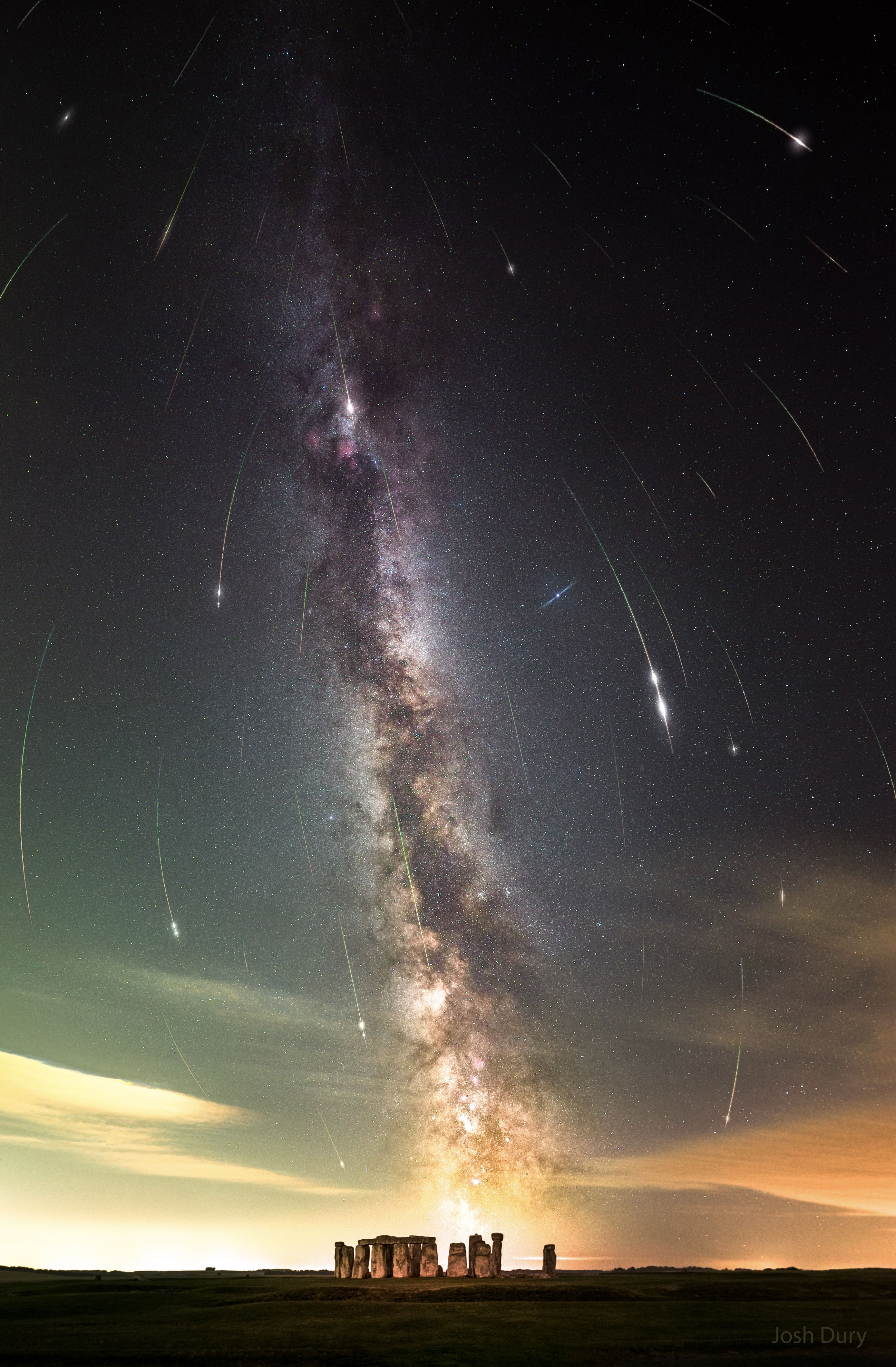 A night sky filled with stars and the band of our Milky Way galaxy is shown also filled with many streaks. The short streaks are all coordinated and together indicate a flow from the top of the image to the bottom. In the foreground at the bottom of the frame is Stonehenge. Please see the explanation for more detailed information.