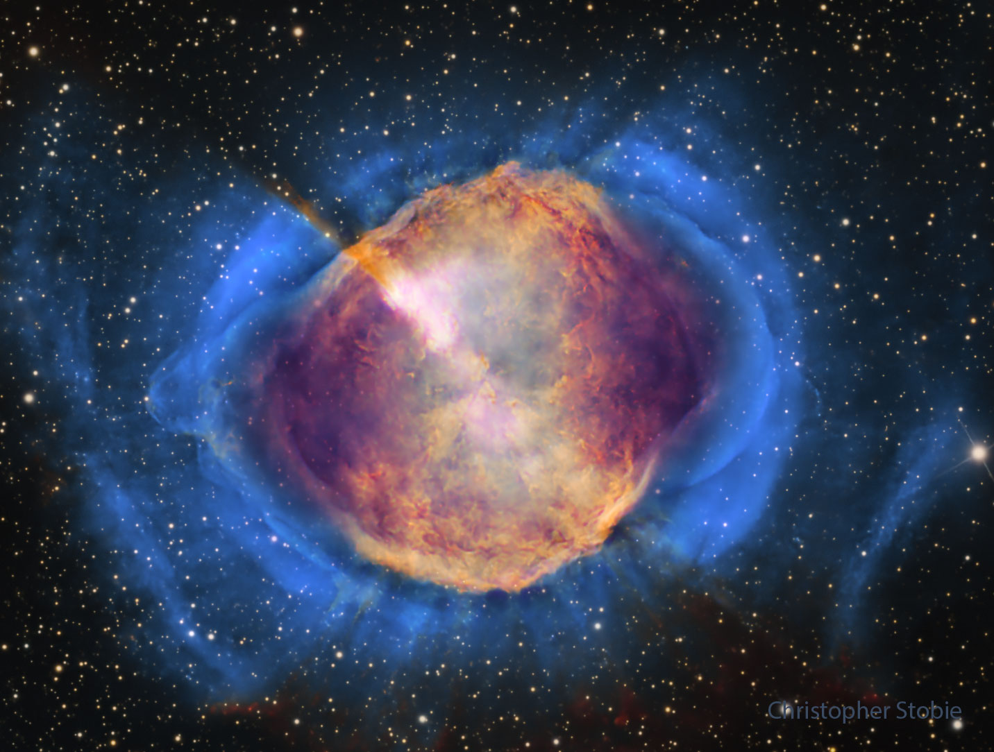 A starfield is shown with a frame dominated by a gaseous nebula. The nebula, filled with structure, appears orange in the center but blue around the edges. Please see the explanation for more detailed information.