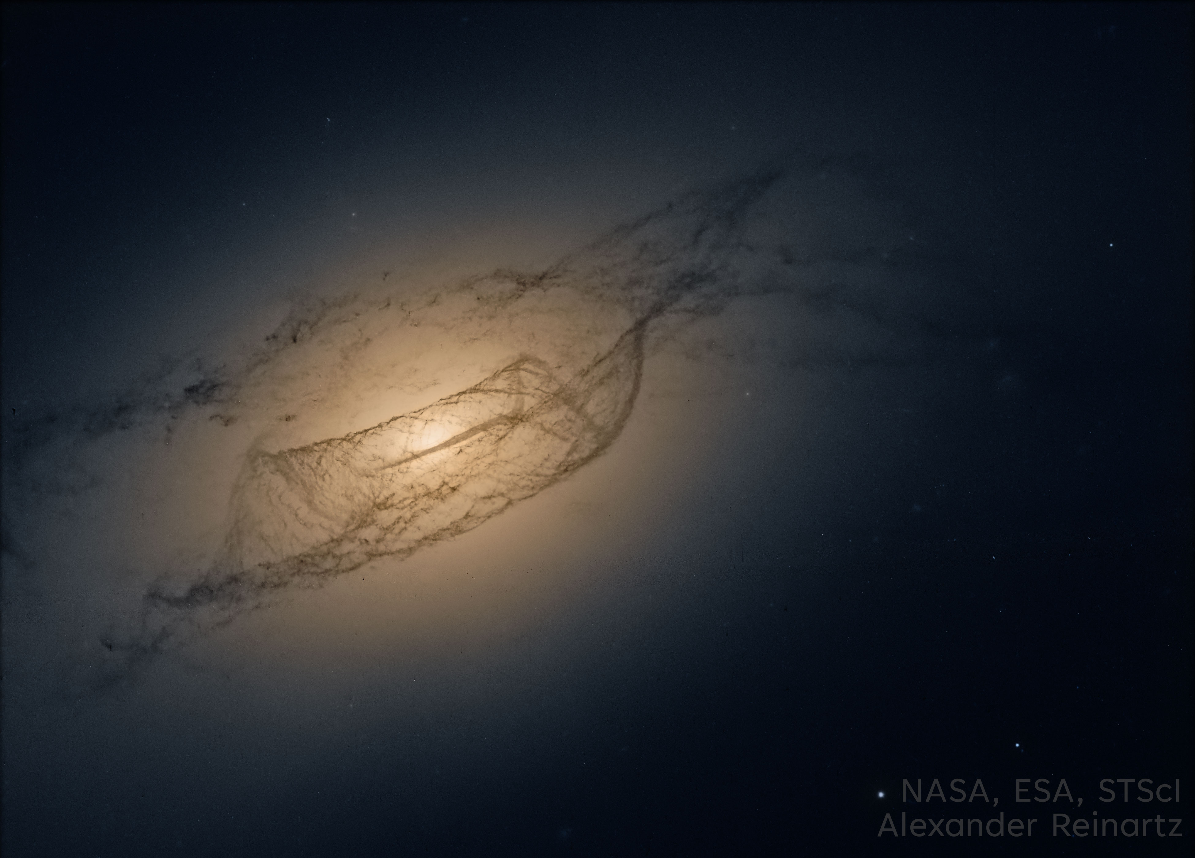 A dark field shows an oblong orange glow with some dark and complex dust lanes running through. Please see the explanation for more detailed information.
