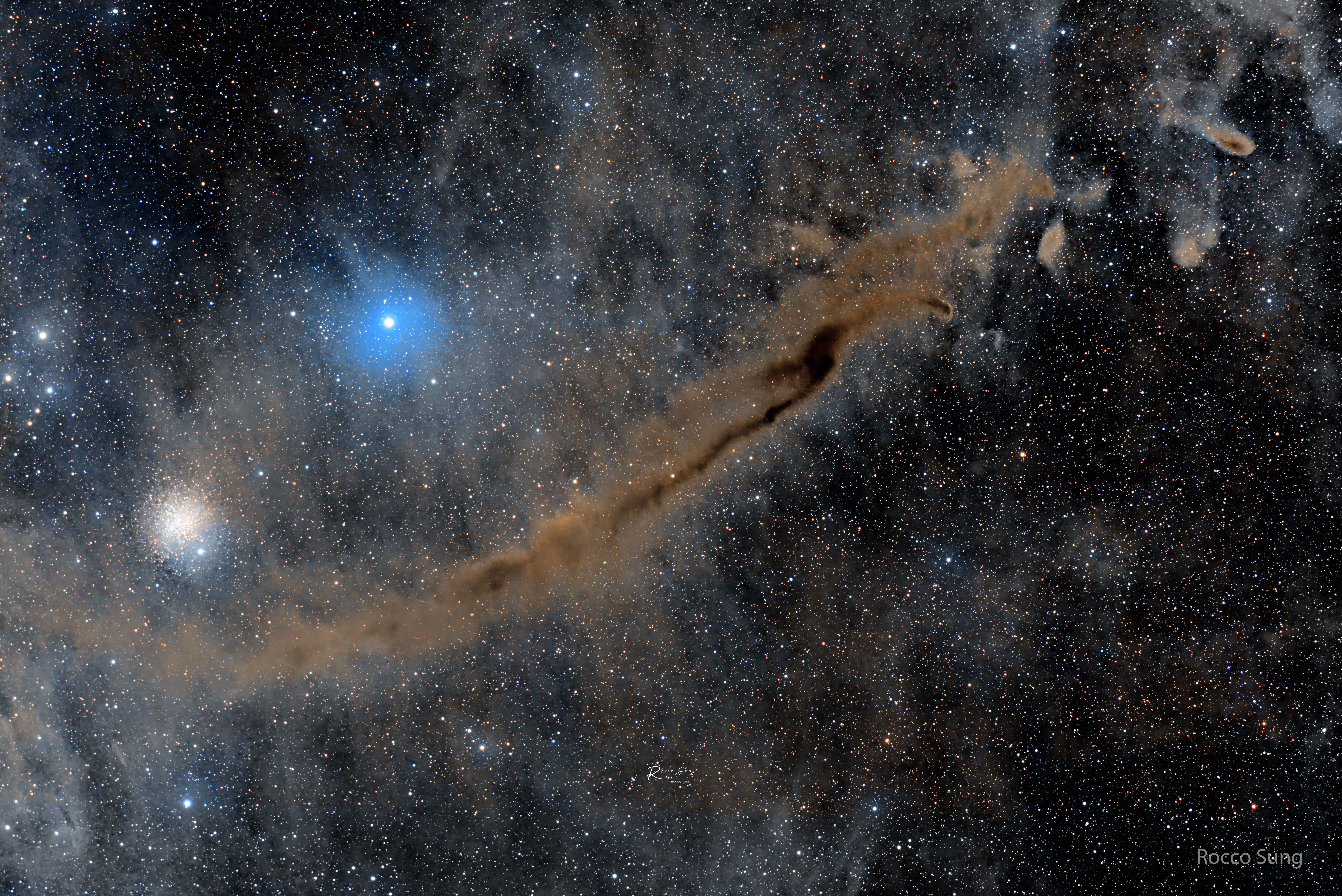 A busy starfield is shown which an elongated brown nebula running diagonally from the lower left to the upper right. A bright blue star and a star cluster appear above the nebula. Please see the explanation for more detailed information.