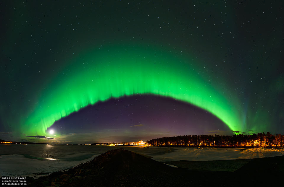 A big green arc is seen arching across the night sky. The arc fades away above into a green haze, while no green glow is seen below the arc. A dark sky filled with stars and constellations fills the background. Snow and distant trees line the foreground. Please see the explanation for more detailed information.