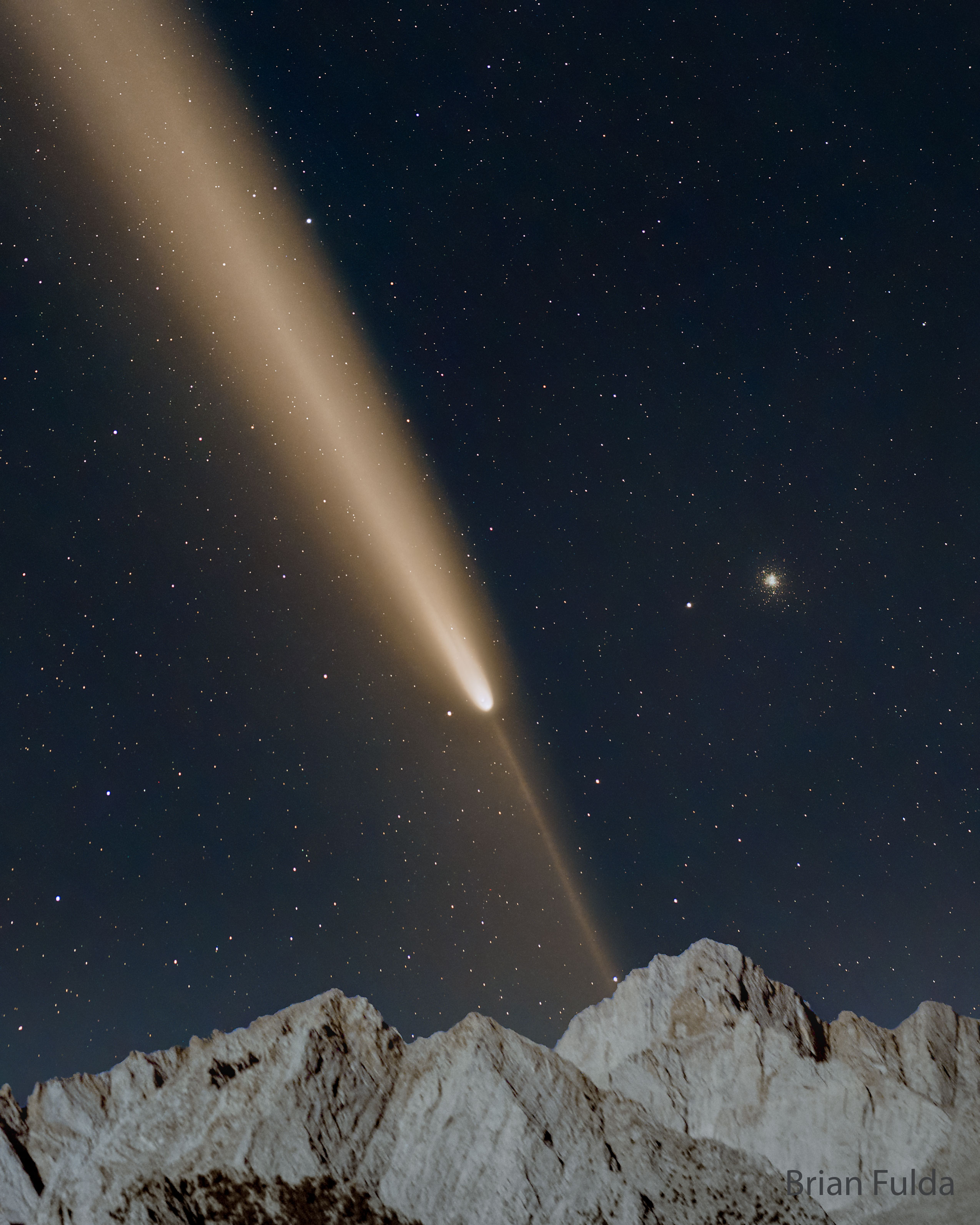A starfield is shown with a bright comet. The main tail of the comet points diagonally to the upper left, while a thin anti-tail points to the lower right. Mountain peaks are visible at the bottom in the foreground. Please see the explanation for more detailed information.