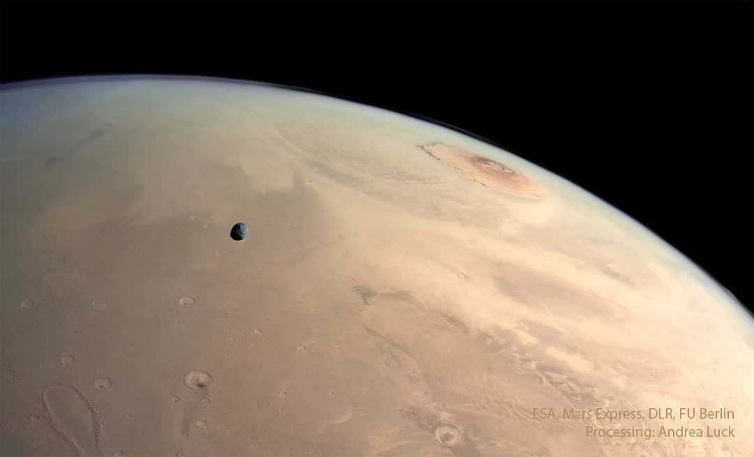 A panoramic view of the surface of Mars. Several landforms are visible including craters and volcanos. A small dark moon is superposed in front of part of the surface. Please see the explanation for more detailed information.