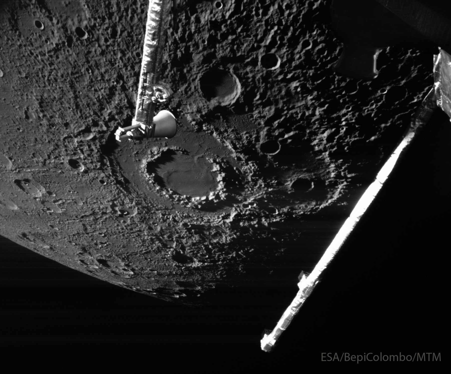 The cratered surface of a large body is shown: Mercury. The largest feature visible is a large impact crater with two rings, near the image center. Arms from the BepiColumbo spacecraft that took the image are seen extending into the image from the top and the right. Please see the explanation for more detailed information.