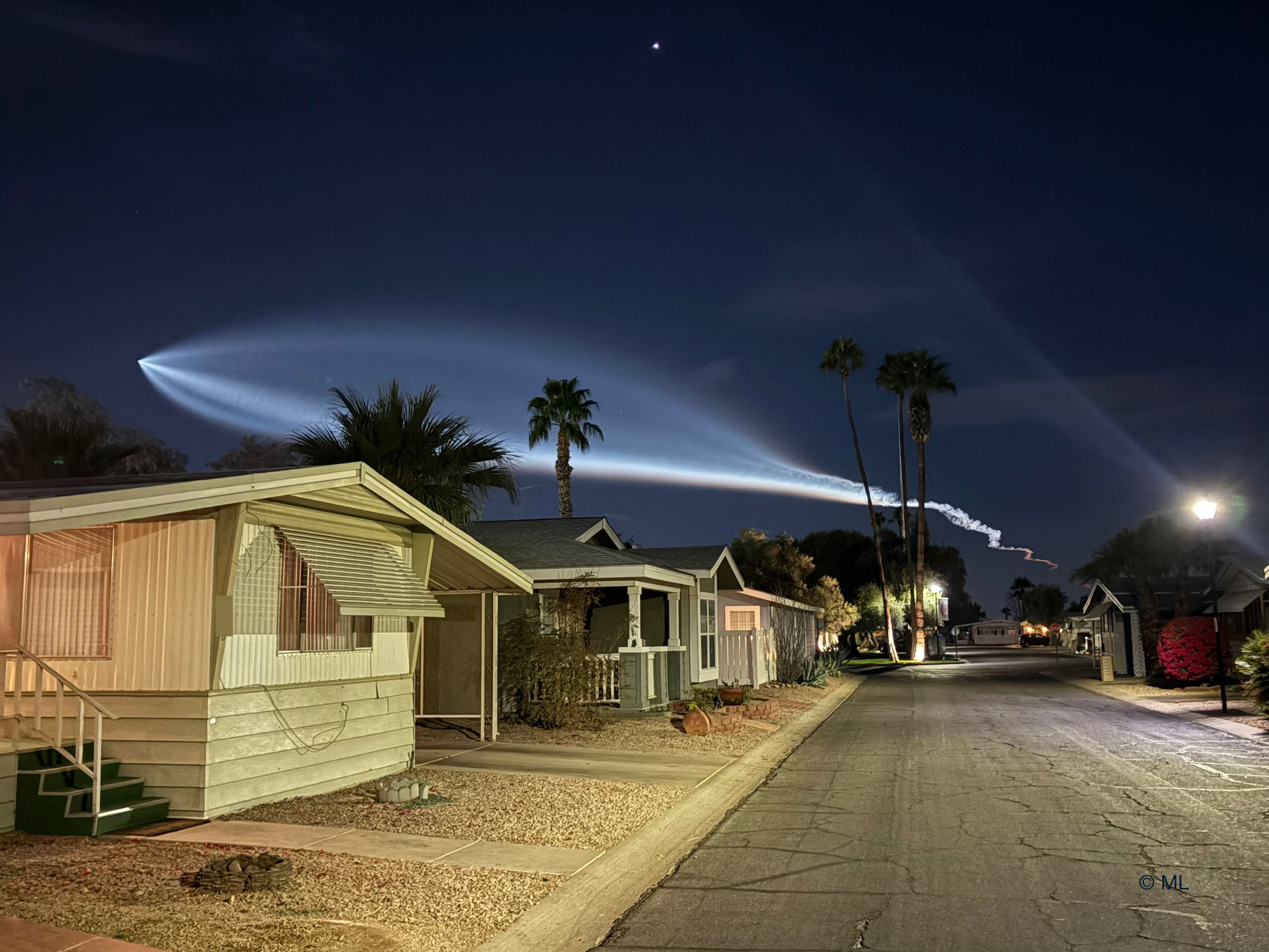 Houses are seen on a street below the night sky. In the sky is a bright light plume that looks like the outline of a giant fish. Please see the explanation for more detailed information.