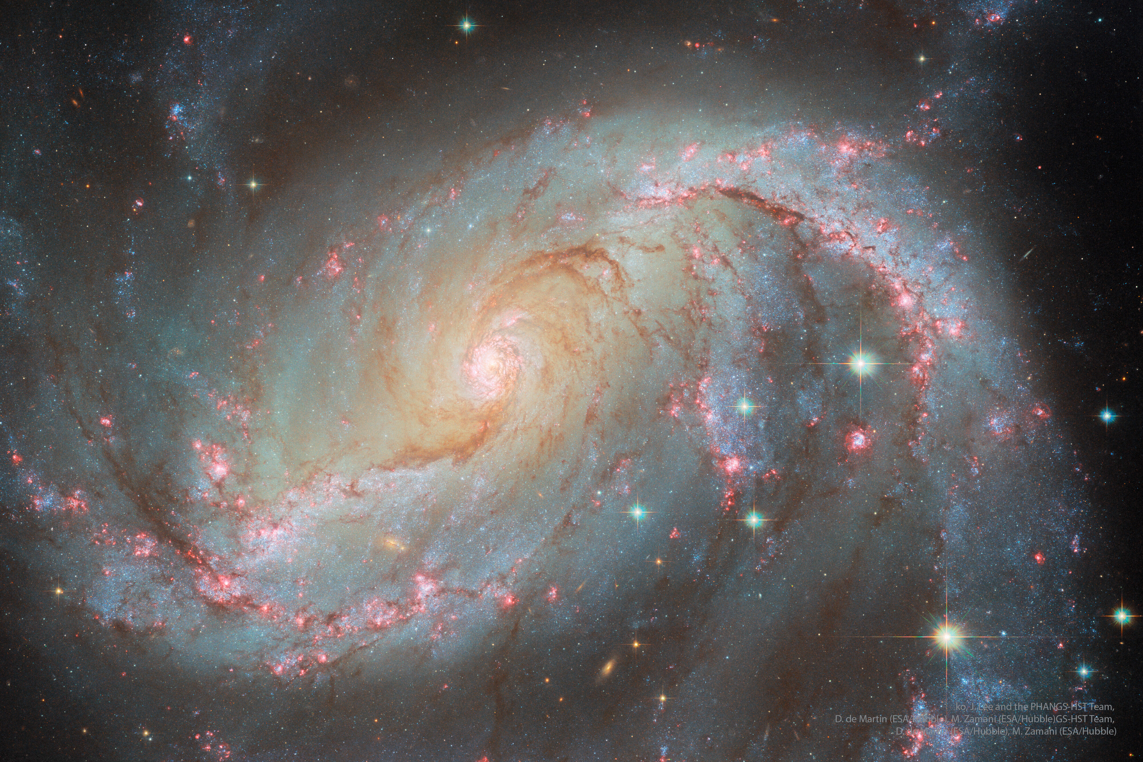 A spiral galaxy is shown in great detail. Visible are blue star clusters, red nebulas, and brown dust in a spiral pattern around the image and galaxy center. Please see the explanation for more detailed information.
