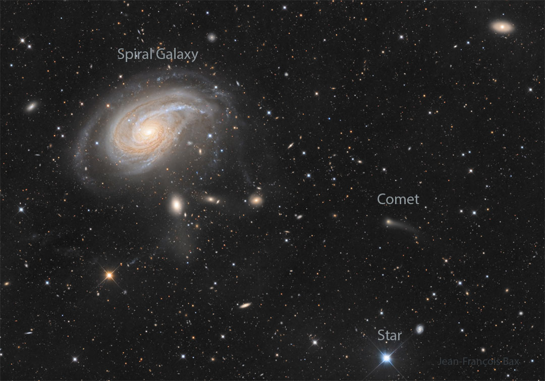 A dark field is filled with stars and galaxies. A large spiral galaxy appears on the upper left. Toward the right, there is a smaller fuzzy patch that is a comet with a short tail. Please see the explanation for more detailed information.