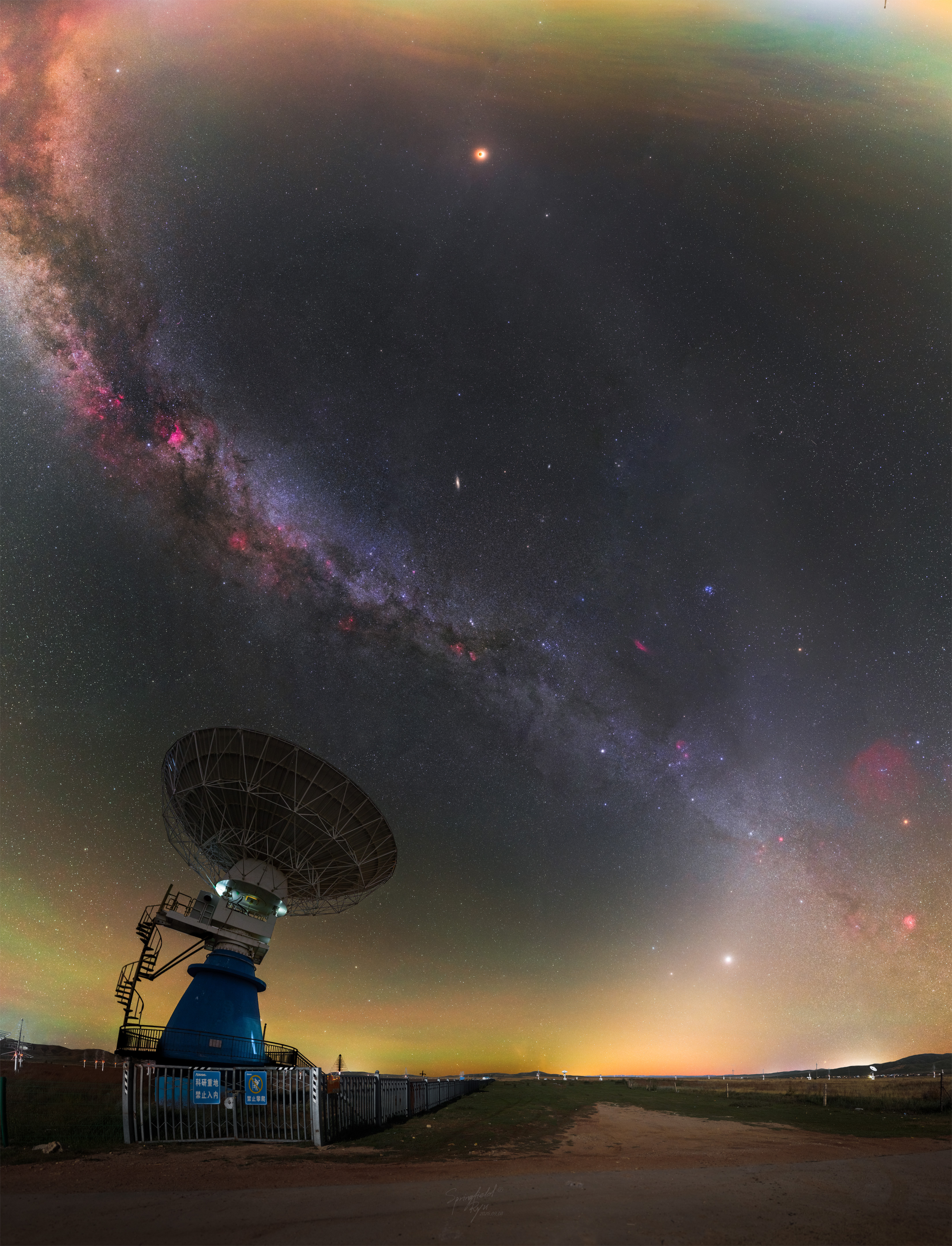 A wide angle image of the night sky is punctuated by a radio telescope on the lower left. Two bands of light run through the sky: the Milky Way and zodiacal light. The bands intertwine like a double helix. Near the top is an orange orb that is a totally eclipsed moon. Please see the explanation for more detailed information.