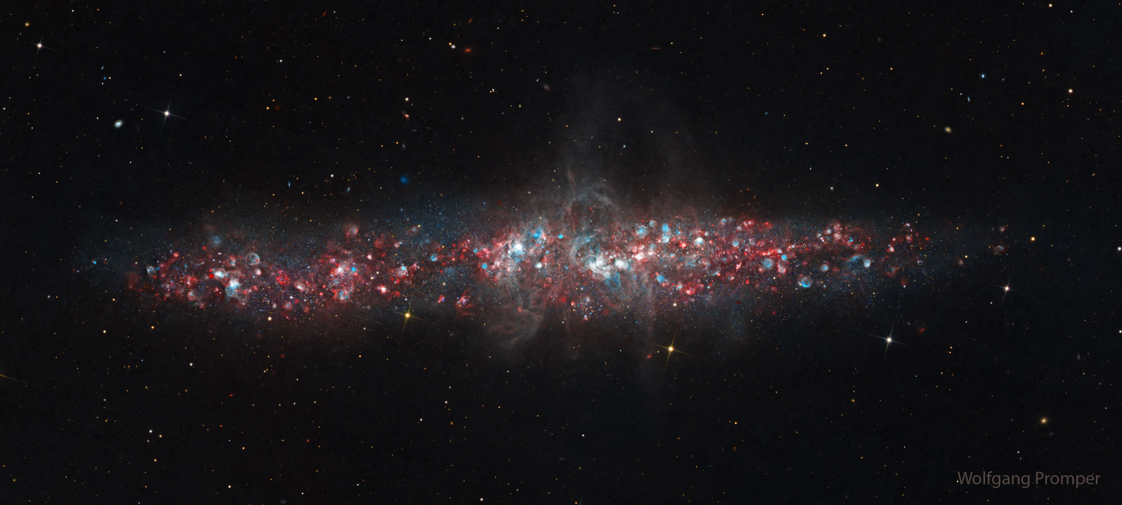 A galaxy is shown that has a lot of blue and red dots in it. The galaxy is an long elliptical blob across the middle and not a classic spiral. Please see the explanation for more detailed information.
