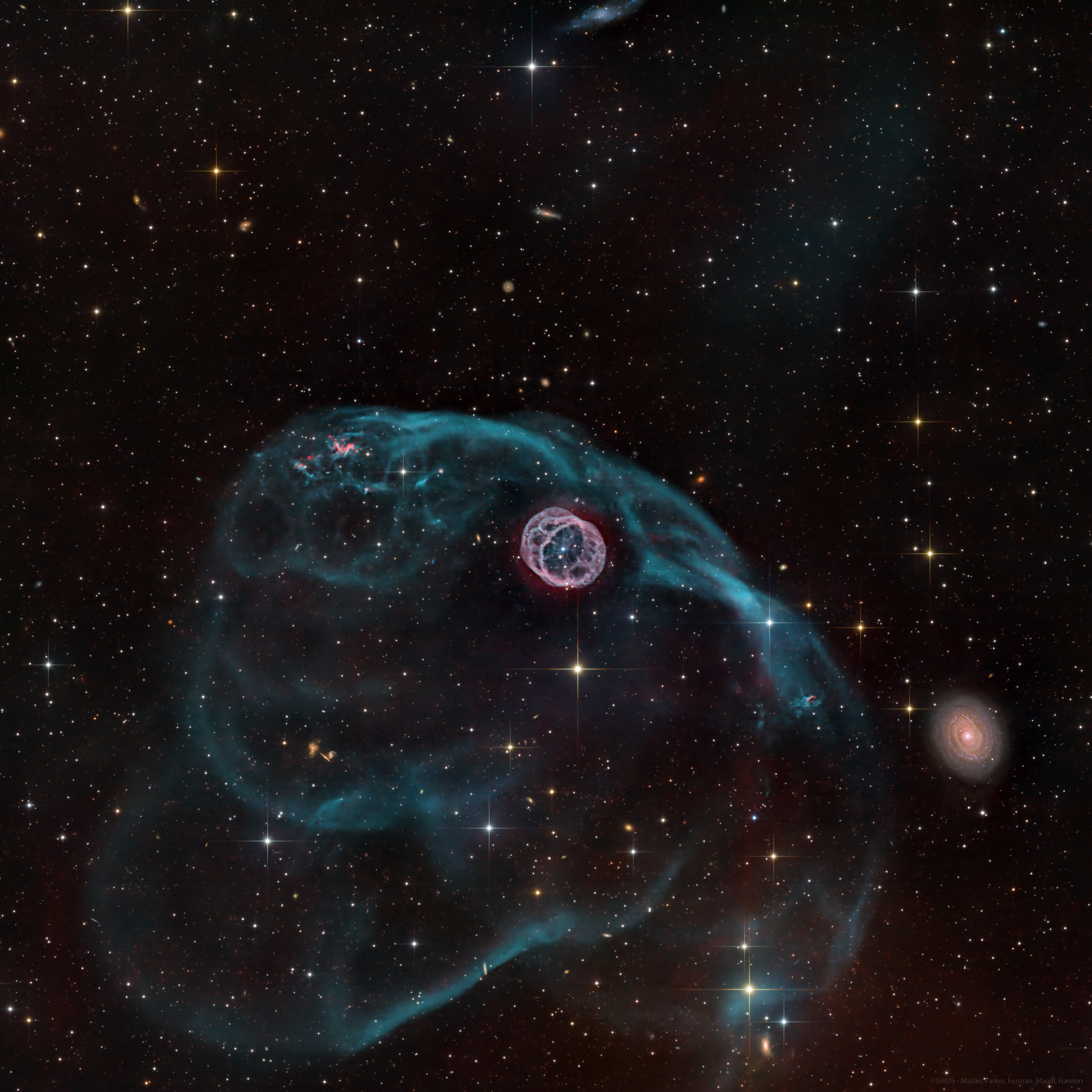 A gaseous structure that resembles a hamster wheel sits near the center of the image. Inside is a bright white dwarf star. There is a larger asymmetric gas bubble surrounding the inner nebula. A galaxy sits to the bottom right. The background is composed of foreground and background stars, as well as distant galaxies.