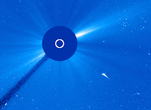 Typical of LASCO C3 data, the image is rendered in blues. A dark �occulting disk,� which is what we call the structure in the telescope that blocks the direct (blinding) sunlight, blocks the Sun. A white circle on the occulting disk indicates the size of the Sun itself. We can see the corona radiating outward in uneven streaks. Stars dot the field behind the sun. A white line representing a comet streaks through space just below and to the right of the Sun. As it reaches and passes behind the occulting disk, a white burst erupts from the other side.