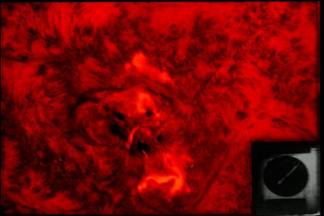 An animated GIF shows a portion of the Sun in red with fuzzy tendrils of gas separated by long, curved dark patches. Near the center, a bright, orange area appears to grow and spread upward, like lava flowing across the Sun's surface, forming a roughly seahorse-shaped feature. A black-and-white inset showing a clock with a hand moving clockwise appears in the lower right.