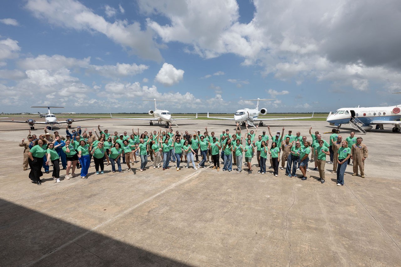 1.	A large group of NASA student airborne research program interns wearing matching bright green t-shirts posing and waving on an airfield tarmac. Several parked airplanes are visible in the background under a sunny, partly cloudy sky.
