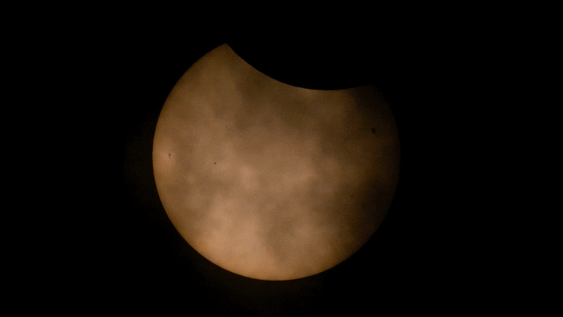 An animated GIF shows the partially eclipsed Sun, with the Moon covering the top part of the Sun and thin clouds in front of the Sun, appearing like smoke. During the animation, a silhouette of the International Space Station passes from right to left across the center of the Sun.
