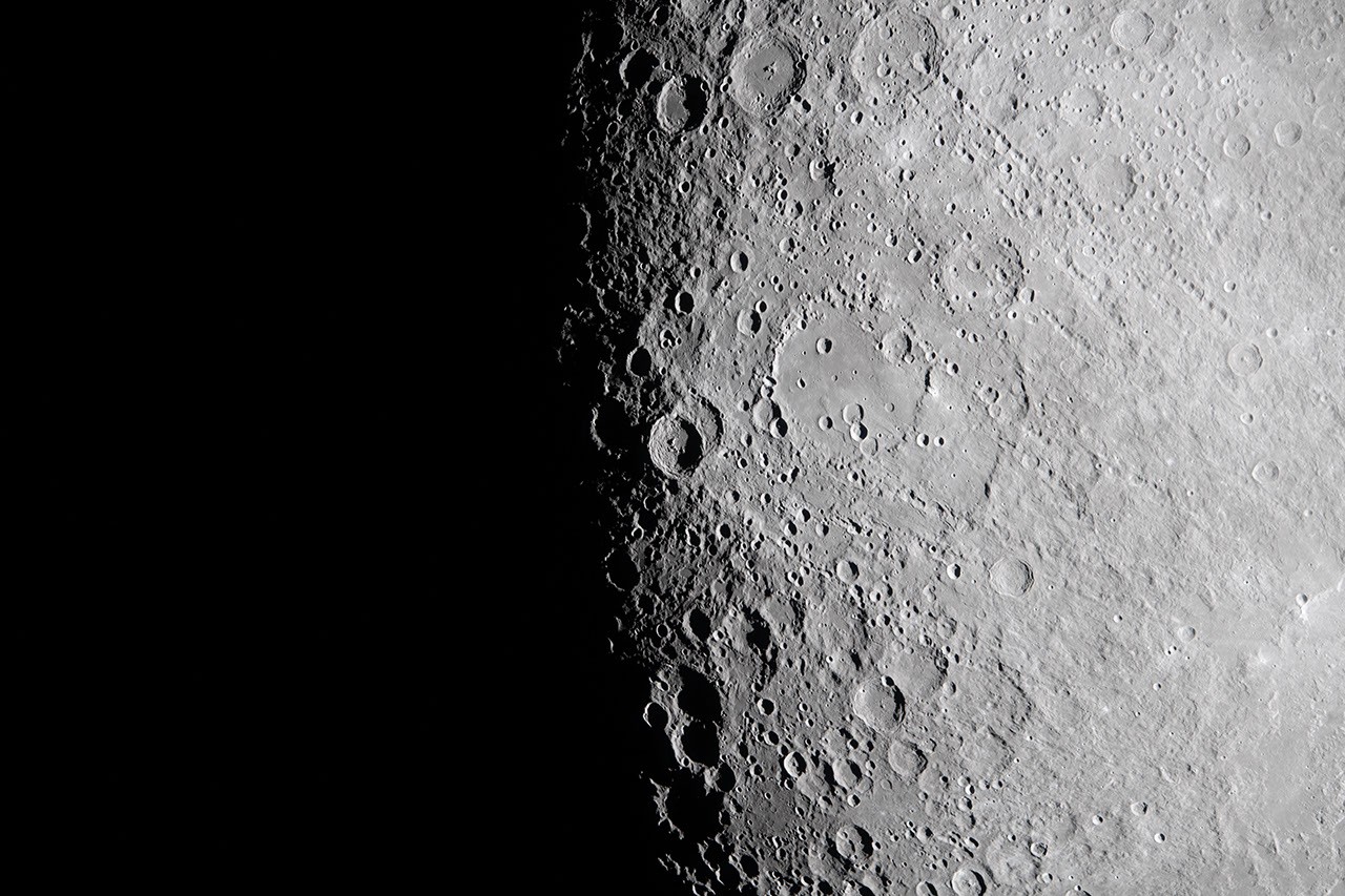 View of the Moon’s surface at the lunar terminator, where bright sunlight meets deep shadow. The right side of the image shows a highly detailed, grey, crater‑covered landscape, with craters of many sizes creating a textured appearance. Toward the left, the surface transitions sharply into complete darkness, marking the boundary between the illuminated and unlit regions of the Moon.